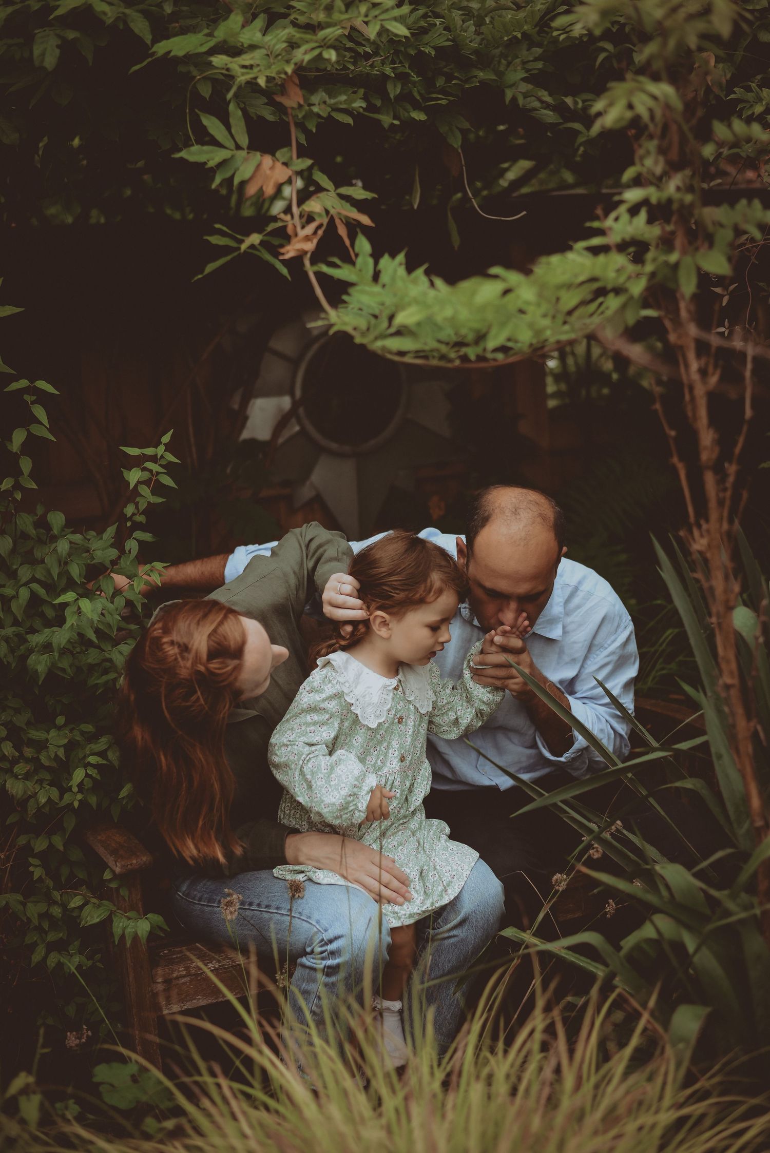 A family of three shares a tender moment surrounded by lush green garden foliage.