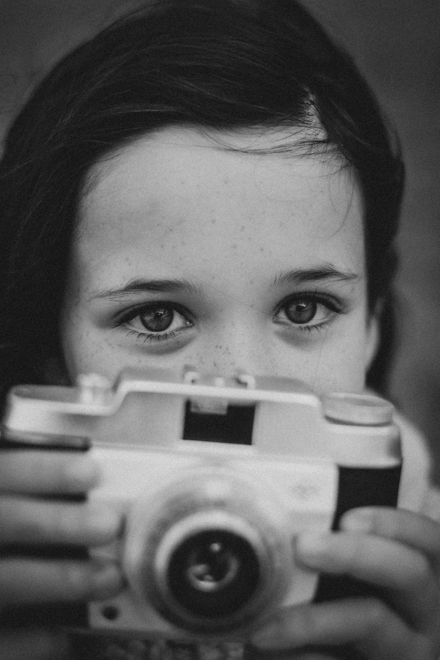 Black and white portrait of a freckled child holding a vintage film camera up to their face.