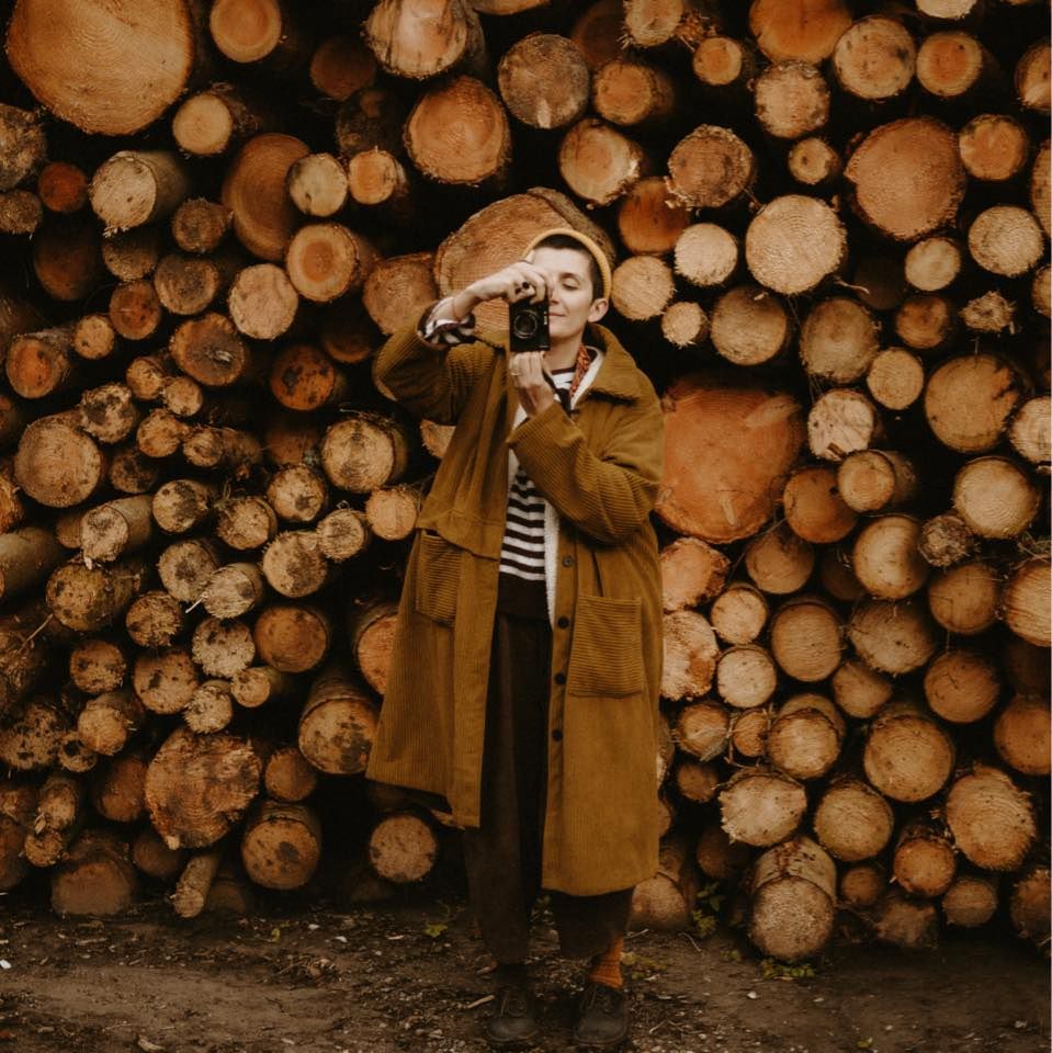 A person in a tan trench coat photographs with a camera in front of stacked logs.