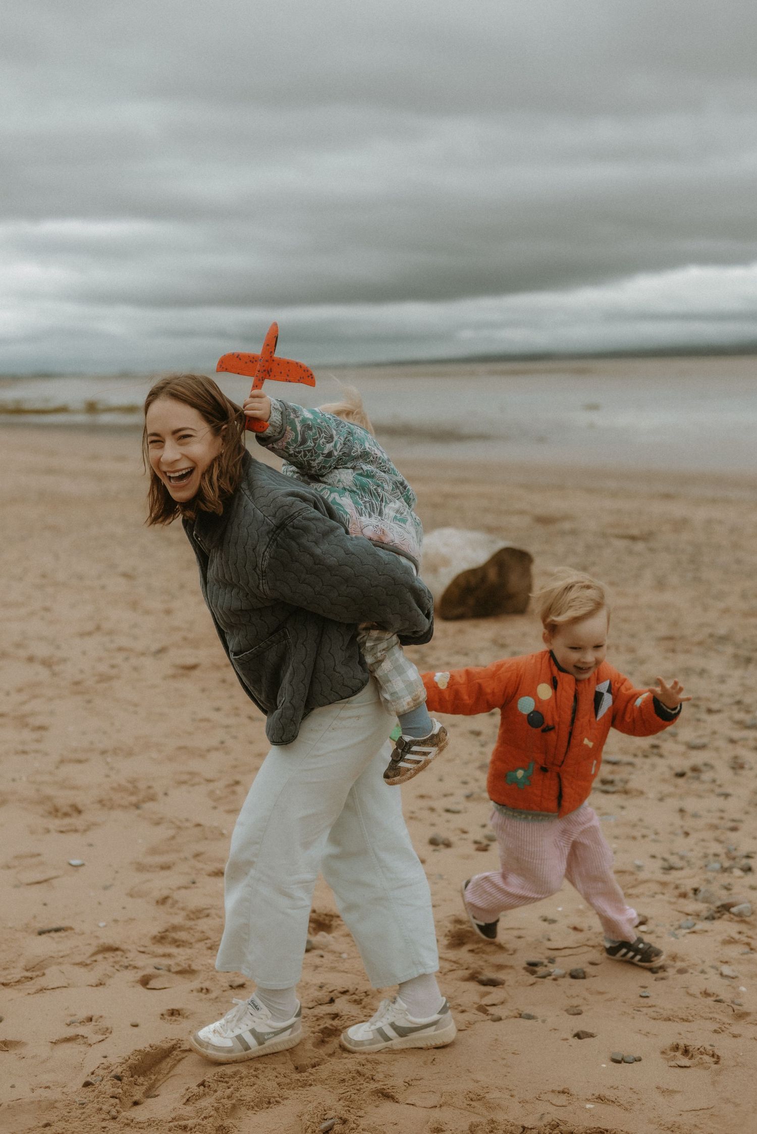A laughing woman carries a child with a toy plane on a sandy beach while a toddler in an orange jacket runs beside her.
