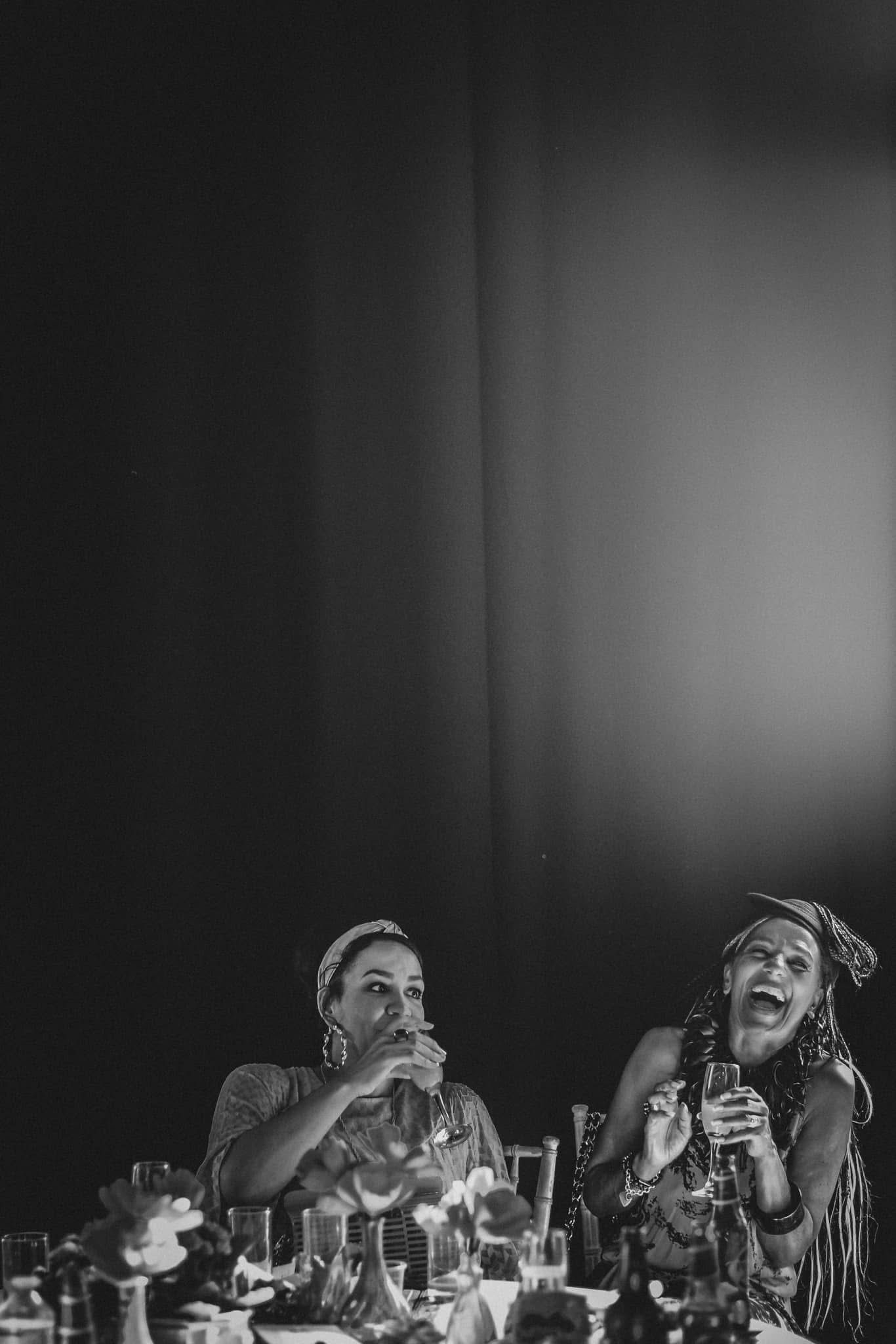 Two women laughing and enjoying drinks at a dinner table, captured in black and white photography.