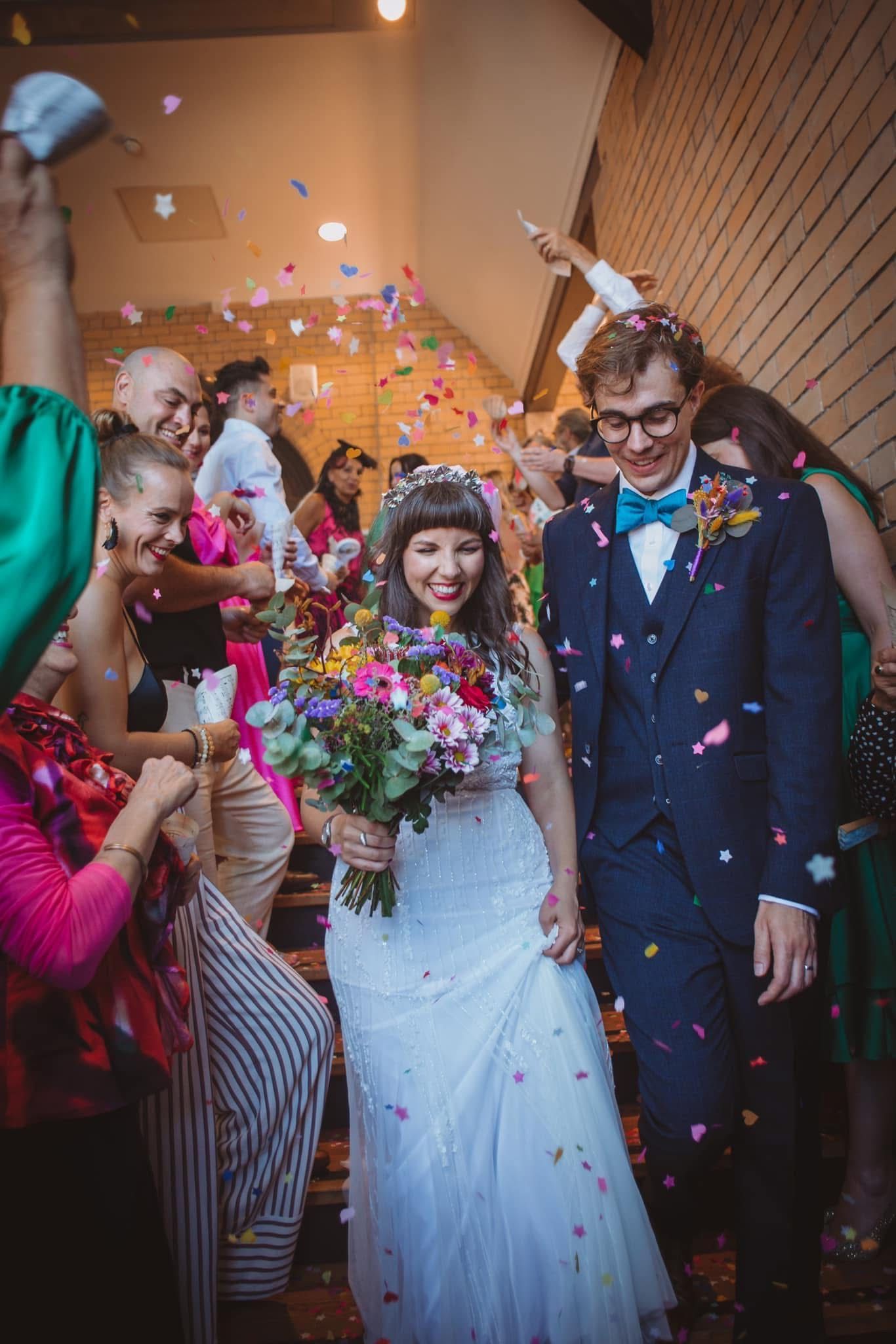Bride and groom walk down the aisle as colorful confetti rains down, surrounded by cheering wedding guests.