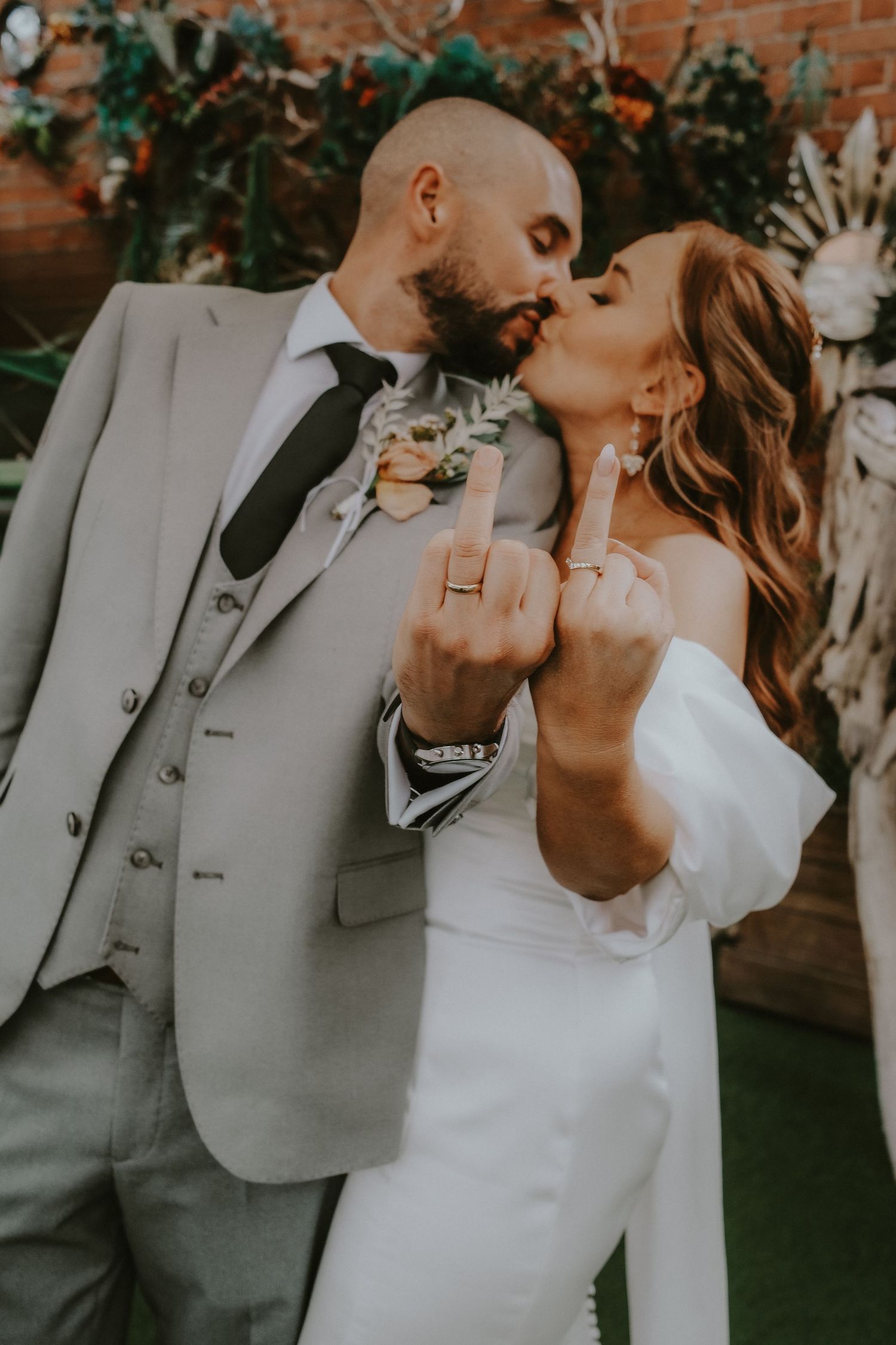 Bride and groom kissing while showing off wedding rings on their fingers outdoors.