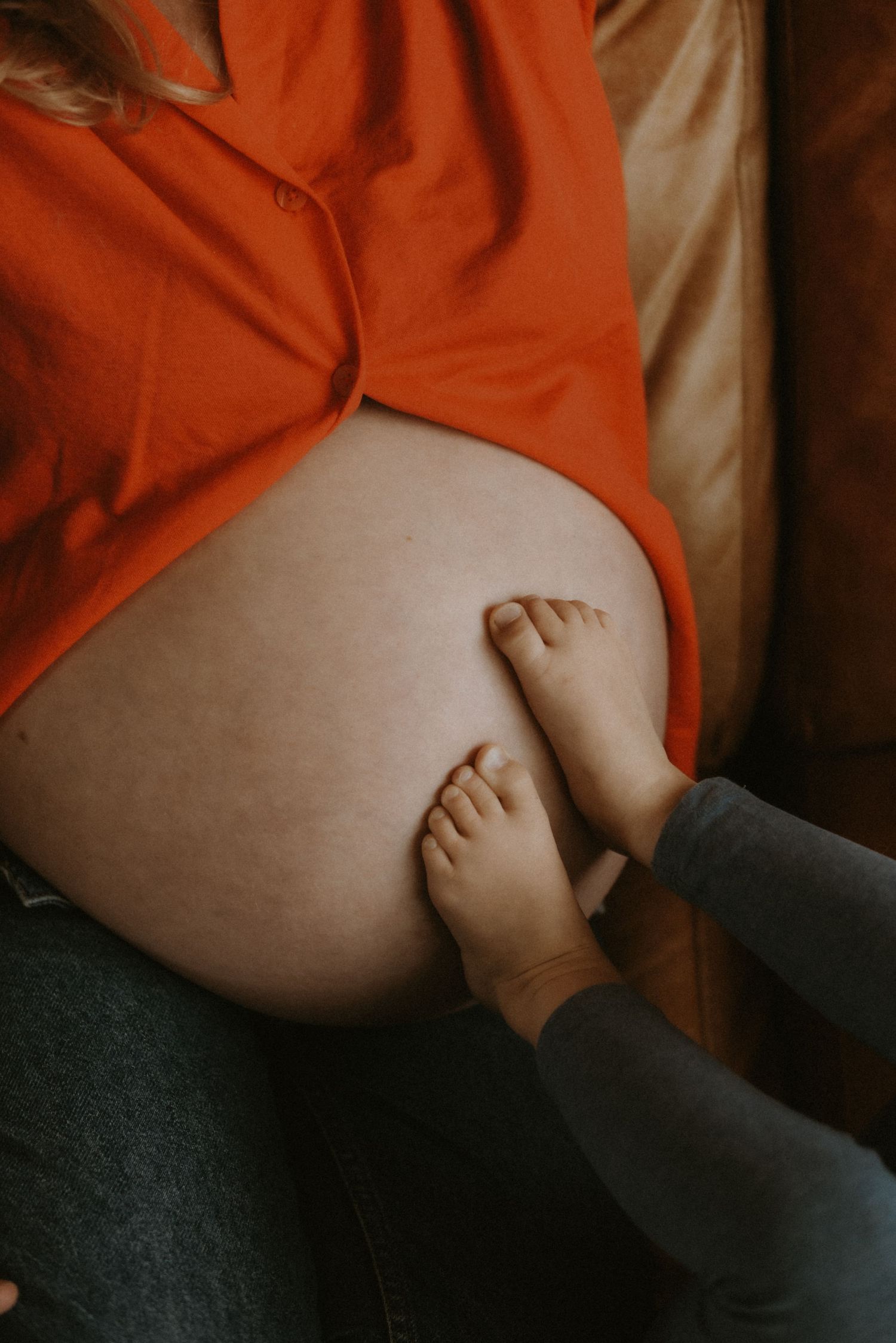 A pregnant woman in an orange shirt sits on a couch while a child's bare feet rest on her baby bump.