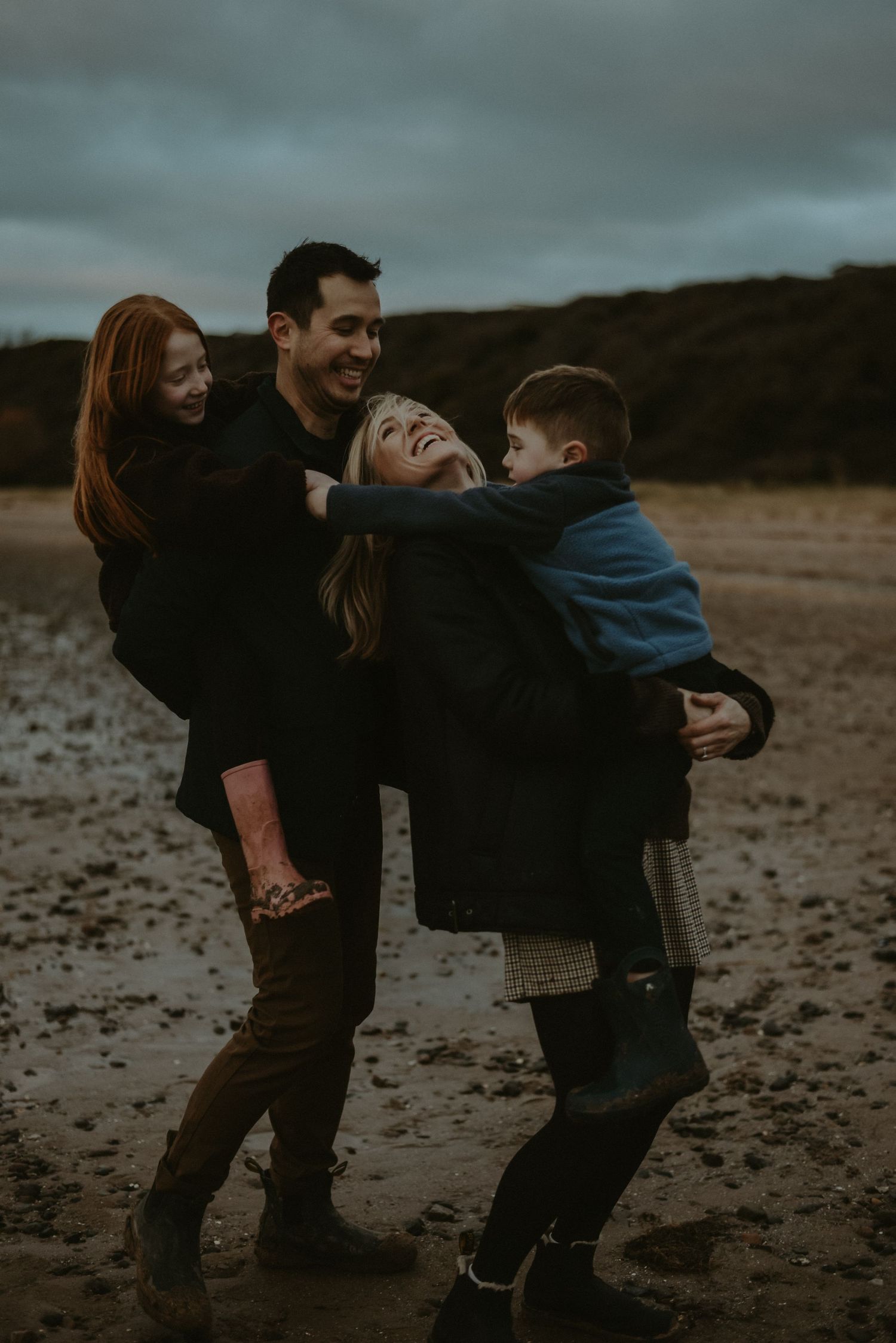 Joyful family of four laughing and embracing on a moody overcast beach at dusk.