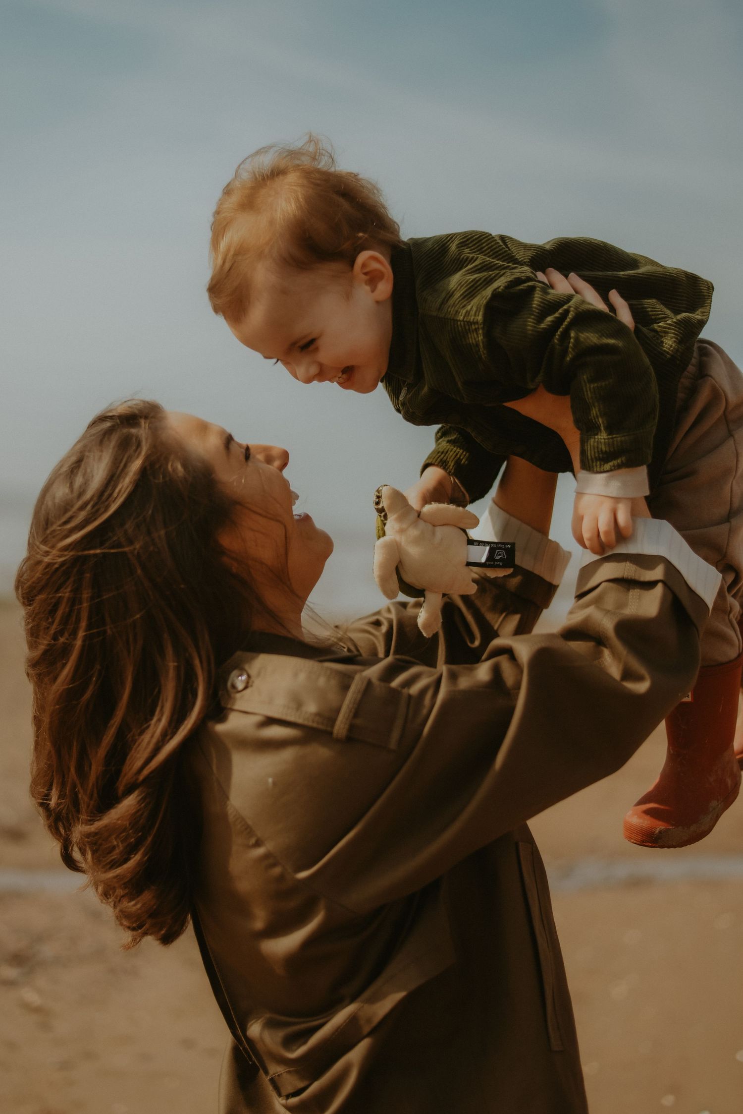 A joyful mother lifts her laughing red-haired toddler high in the air on a sunny beach.