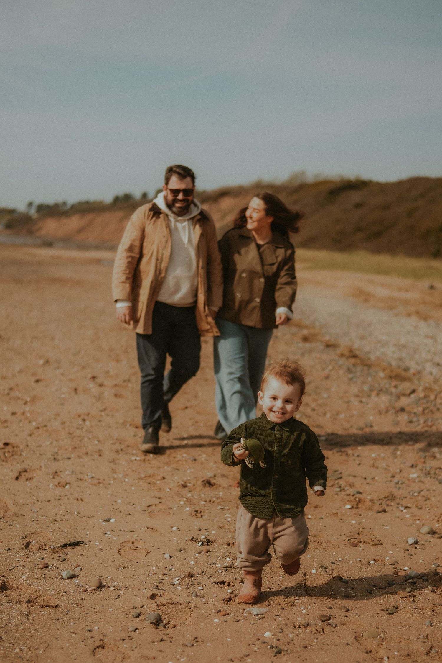 Happy young toddler runs ahead of his smiling parents on a sandy outdoor path in autumn.