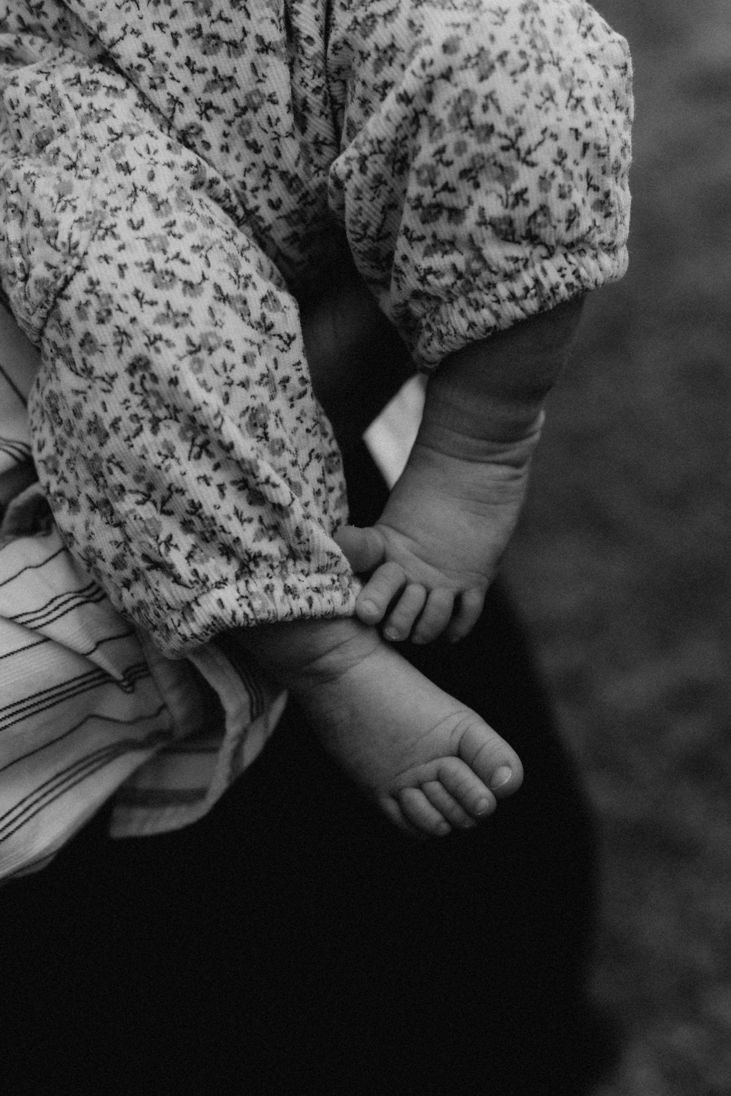 Black and white photo of a baby's tiny feet and legs in a floral onesie, cradled in an adult's arms.