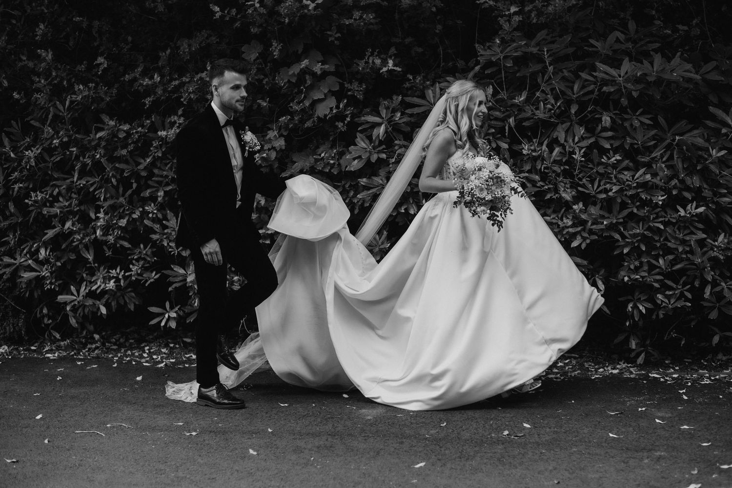Black and white photo of bride and groom walking hand in hand, bride lifting her flowing wedding gown outdoors.