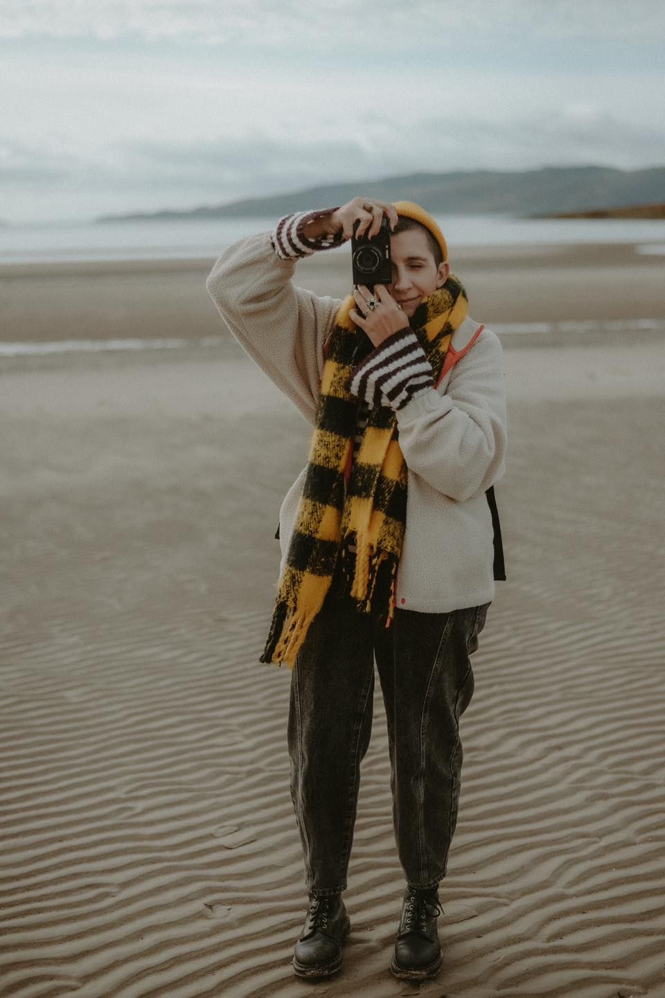 A young person on a sandy beach wearing a yellow scarf and holding a camera.