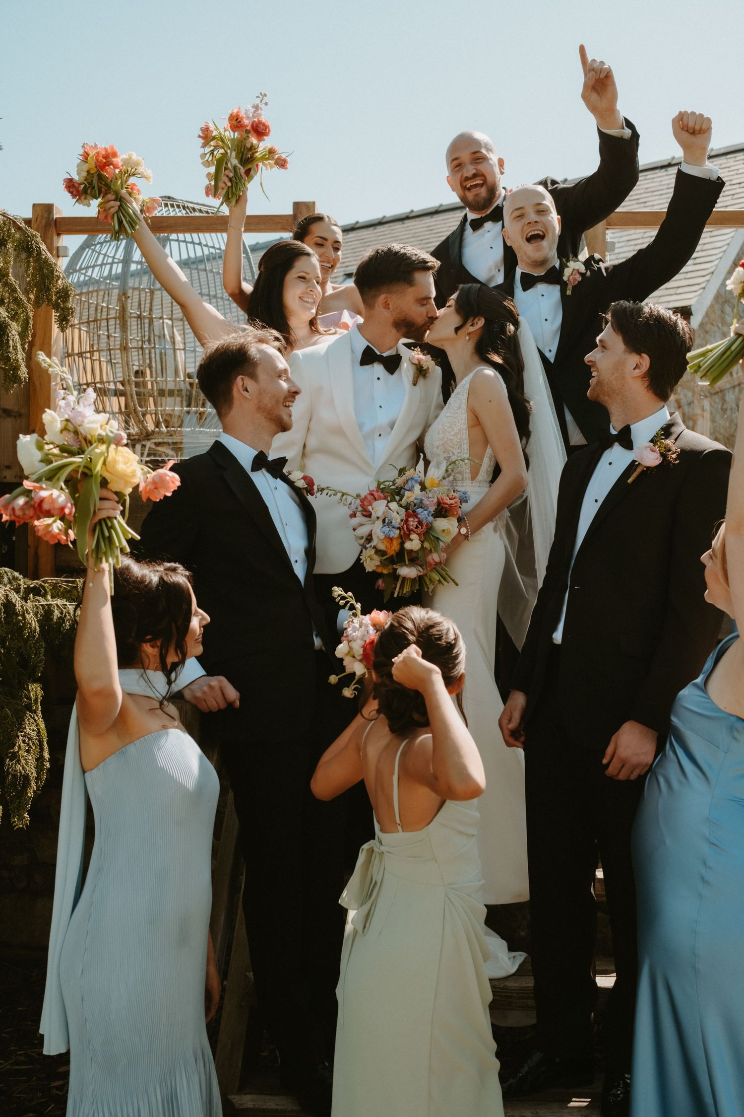 Joyful wedding party celebrating with bride and groom kissing, groomsmen in black tuxedos, bridesmaids holding colorful bouquets.