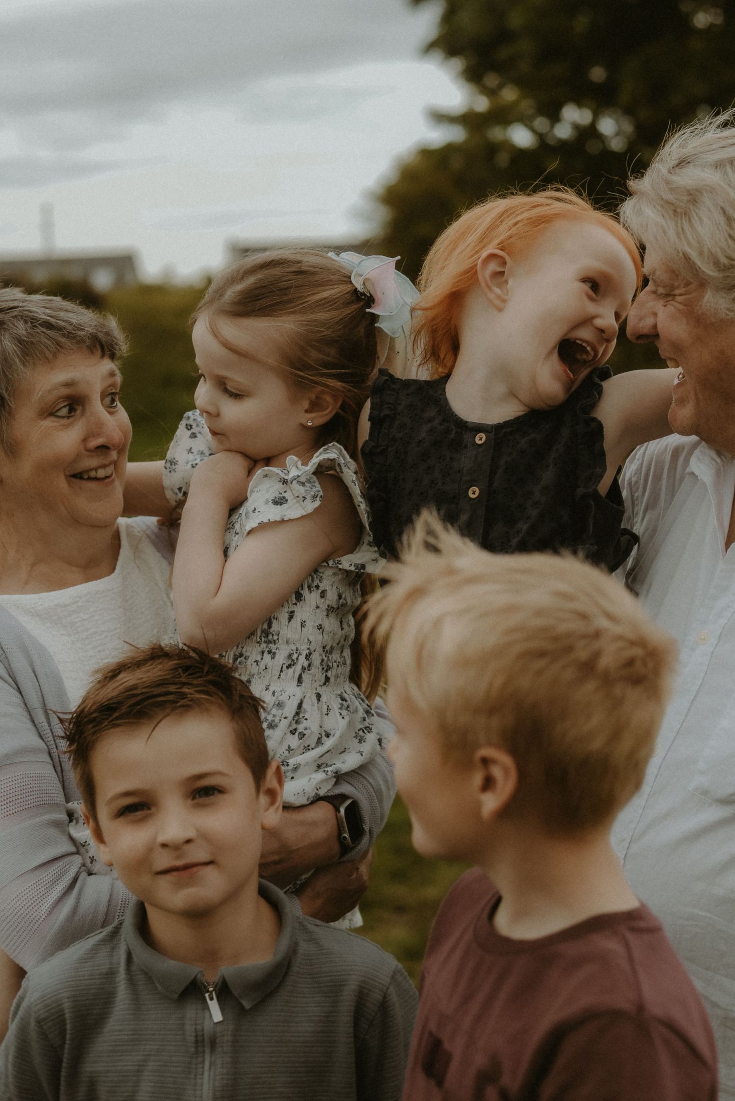 Grandparents laughing and holding young children outdoors with two boys standing in front during a family photo session.