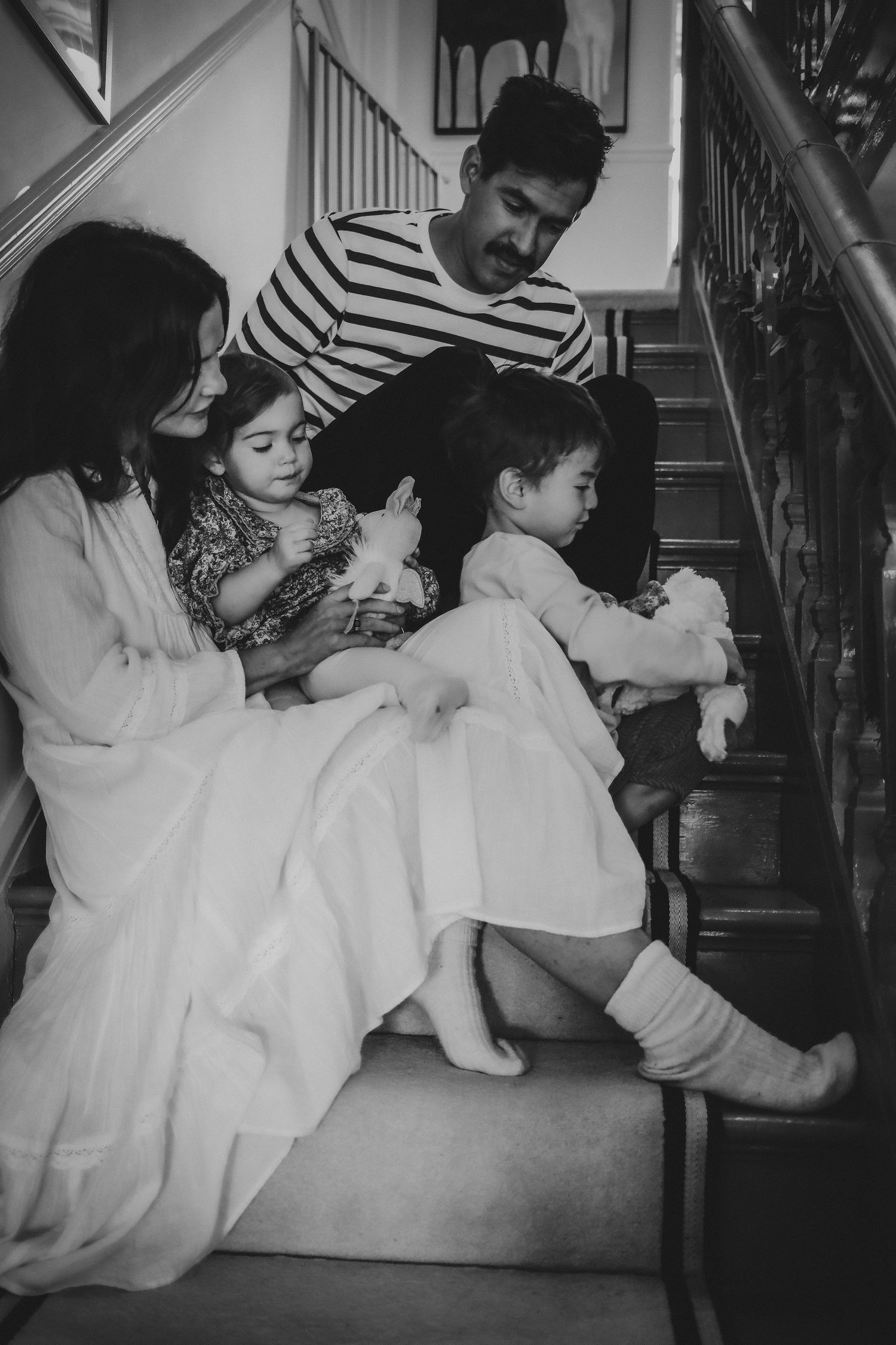 Black and white family portrait on stairs with parents and two young children holding dolls.