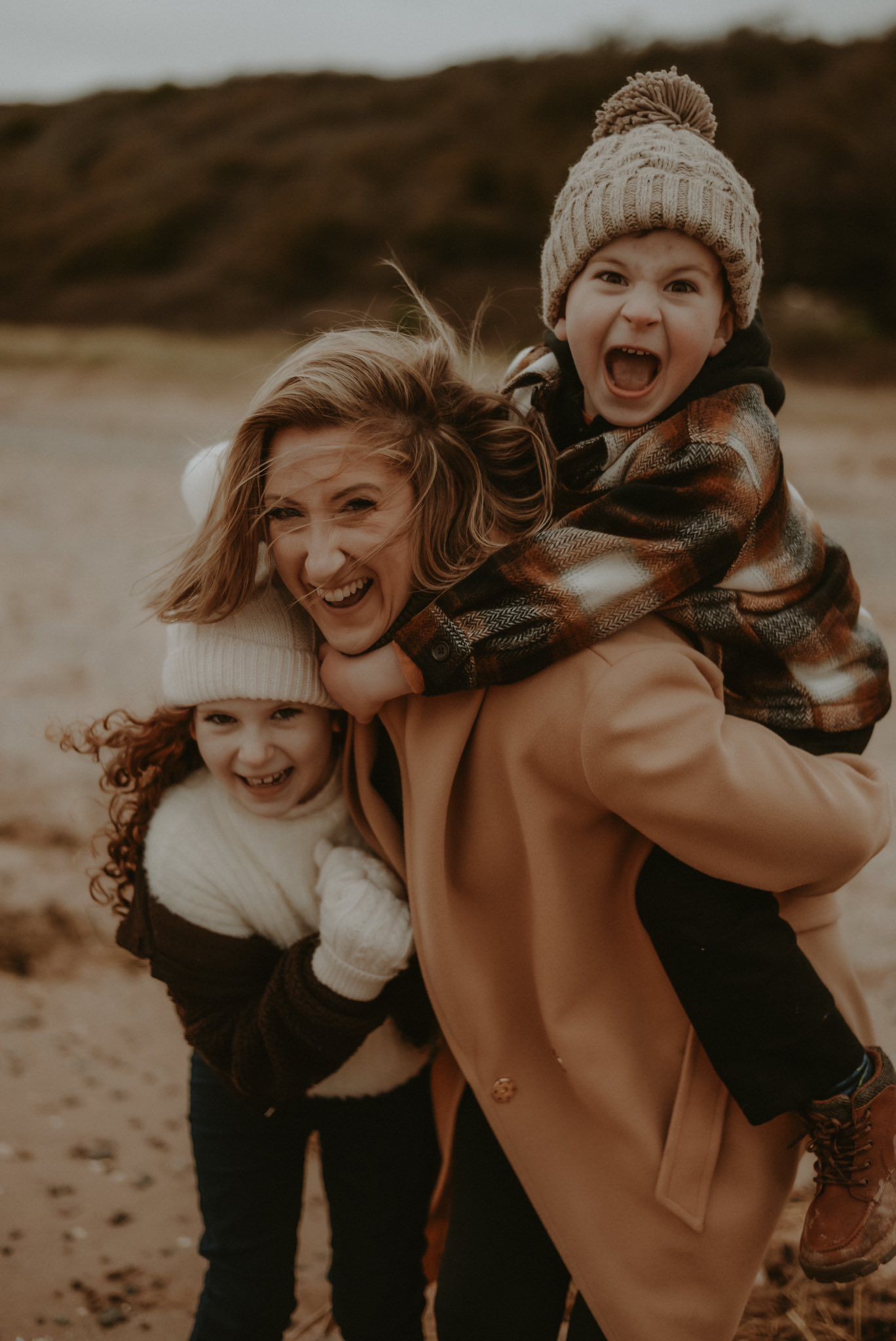 Joyful mother carries two laughing children on a windy beach in warm autumn coats.