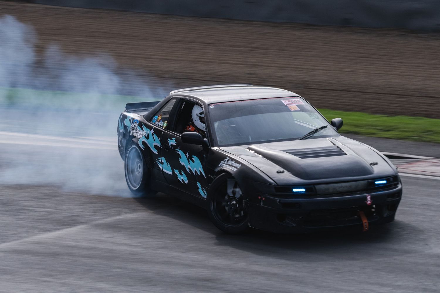 Black drift car with blue flame graphics producing smoke while drifting on a race track.