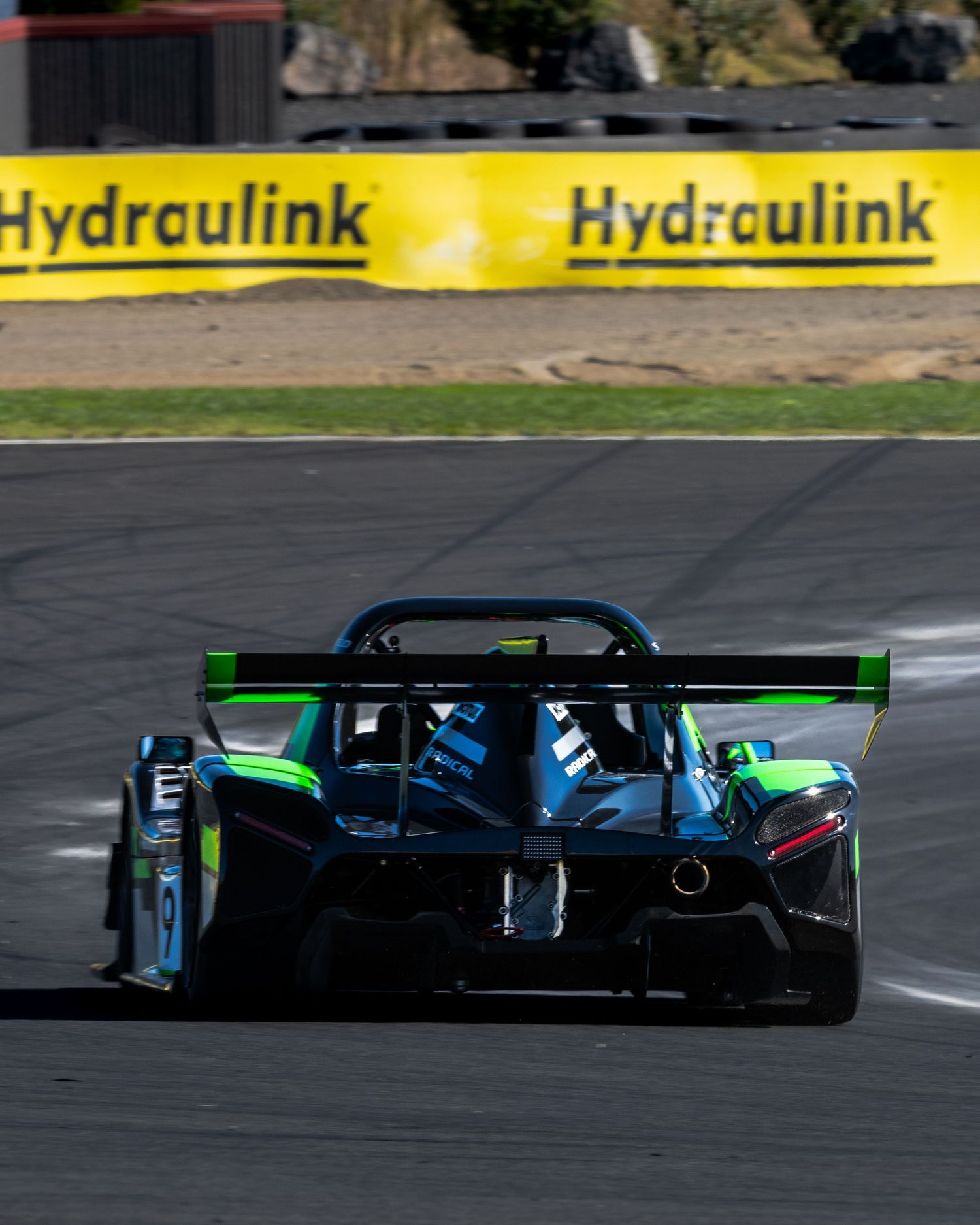 Green and black Radical prototype race car viewed from rear on circuit with Hydraulink signage.