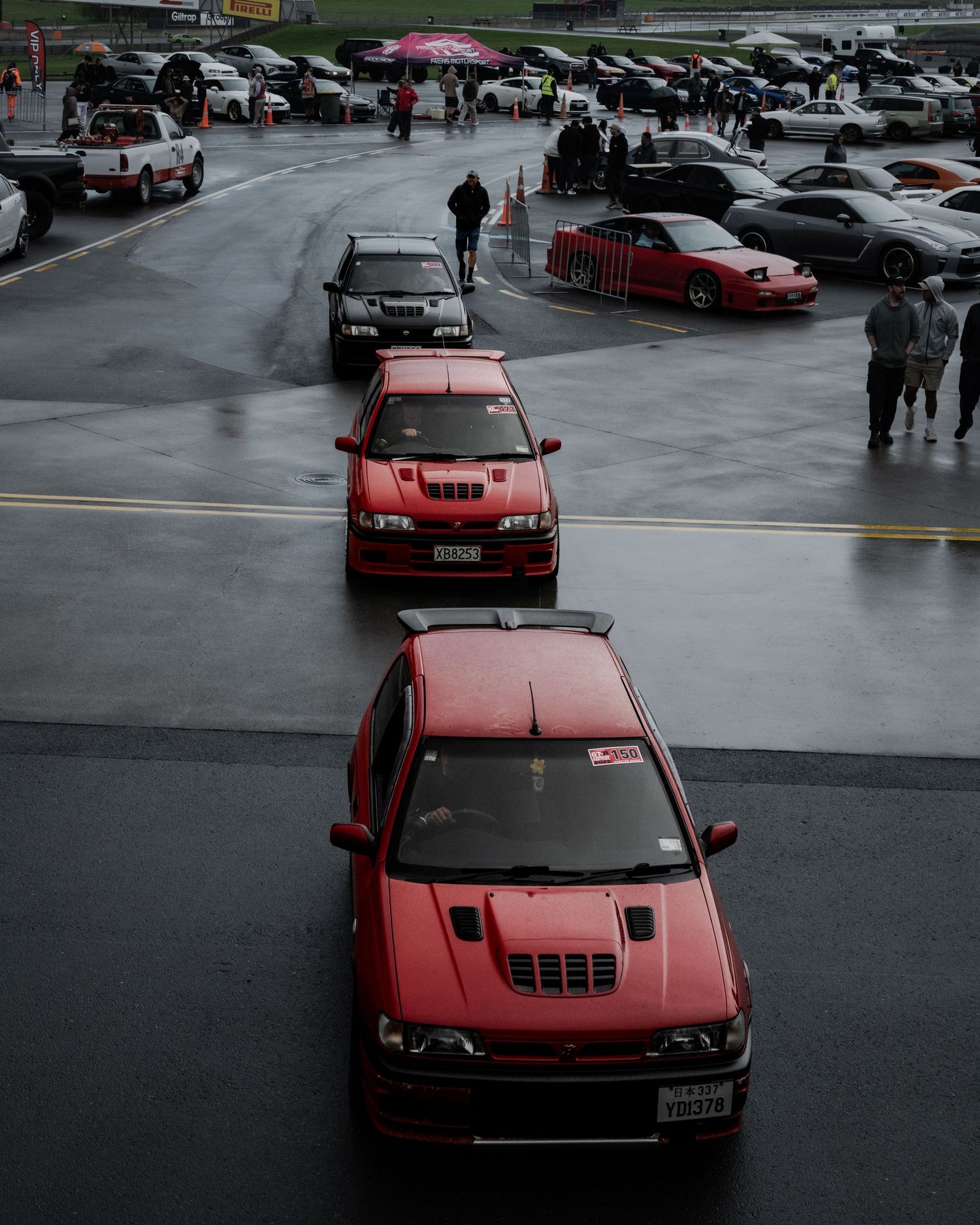 Red Nissan Pulsar GTI-R and other cars queued in a wet racetrack paddock area.