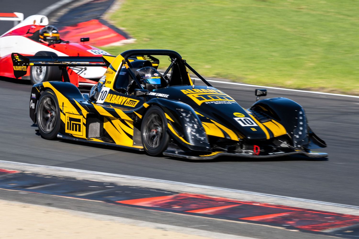Yellow and black Radical race car competing on a circuit track with red car following closely.