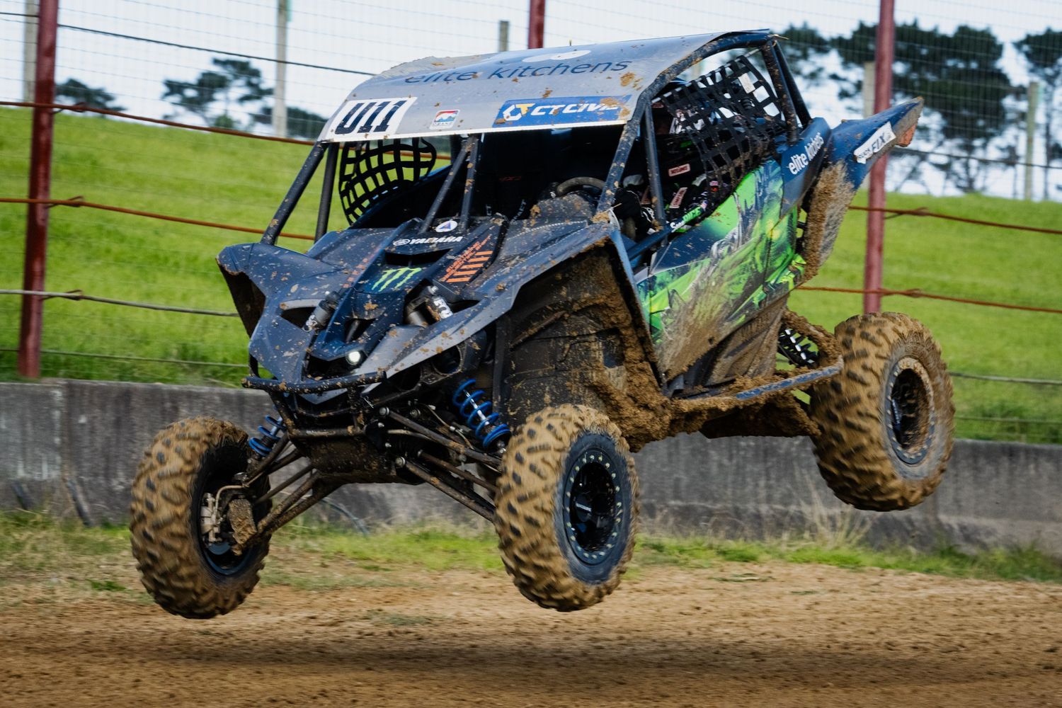 Blue and green UTV side-by-side vehicle airborne during an off-road dirt track race.