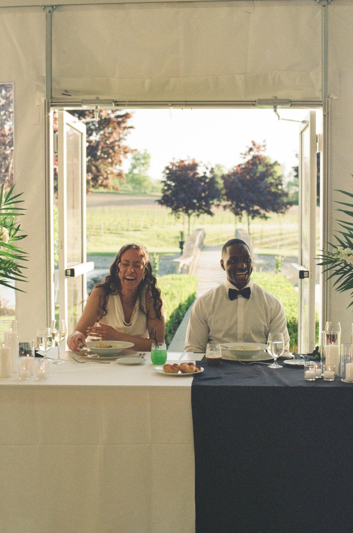 Happy couple laughing at sweetheart table during outdoor wedding reception under white tent.