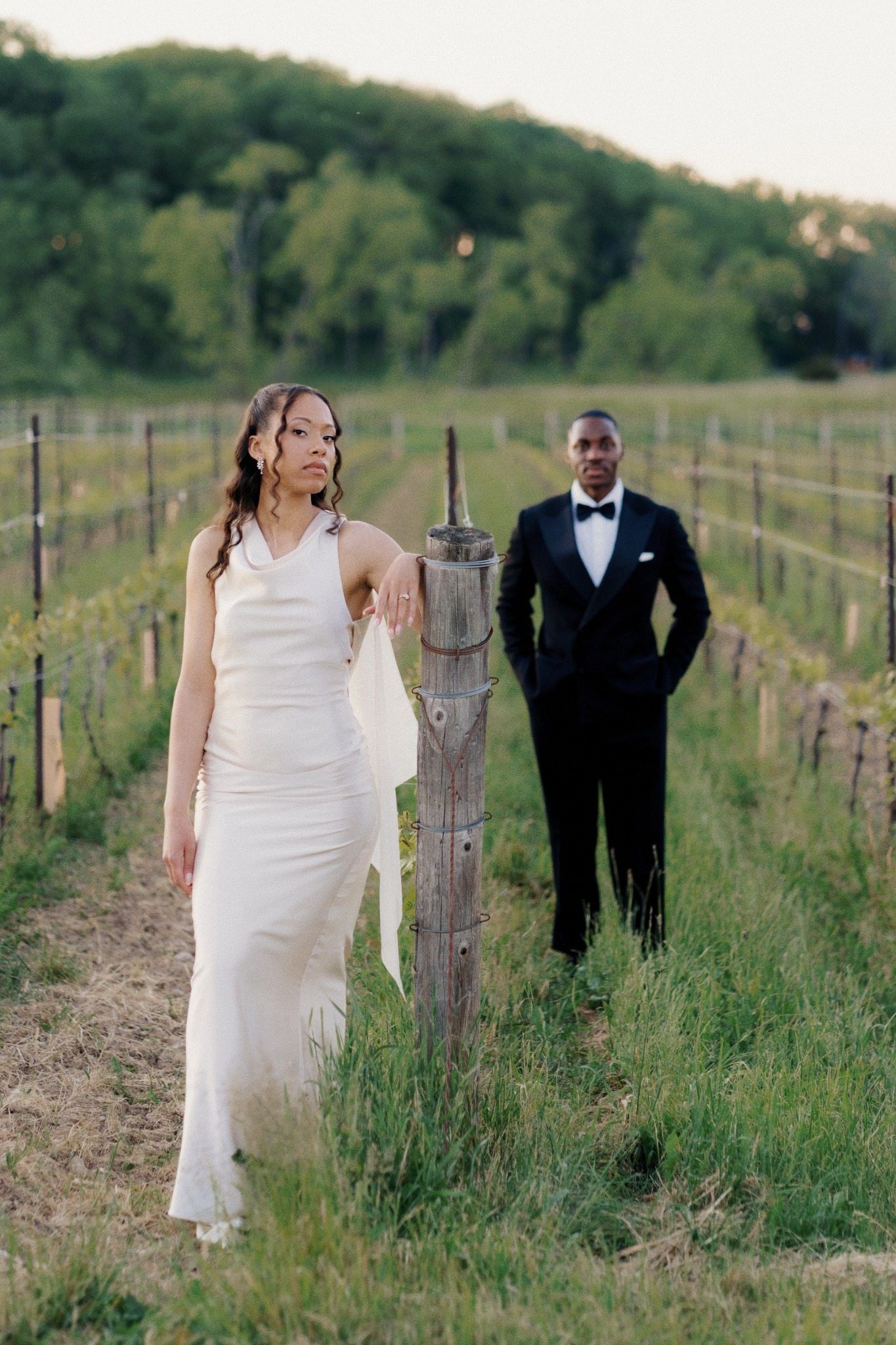 Bride in white gown and groom in black tuxedo pose in a lush vineyard at golden hour.