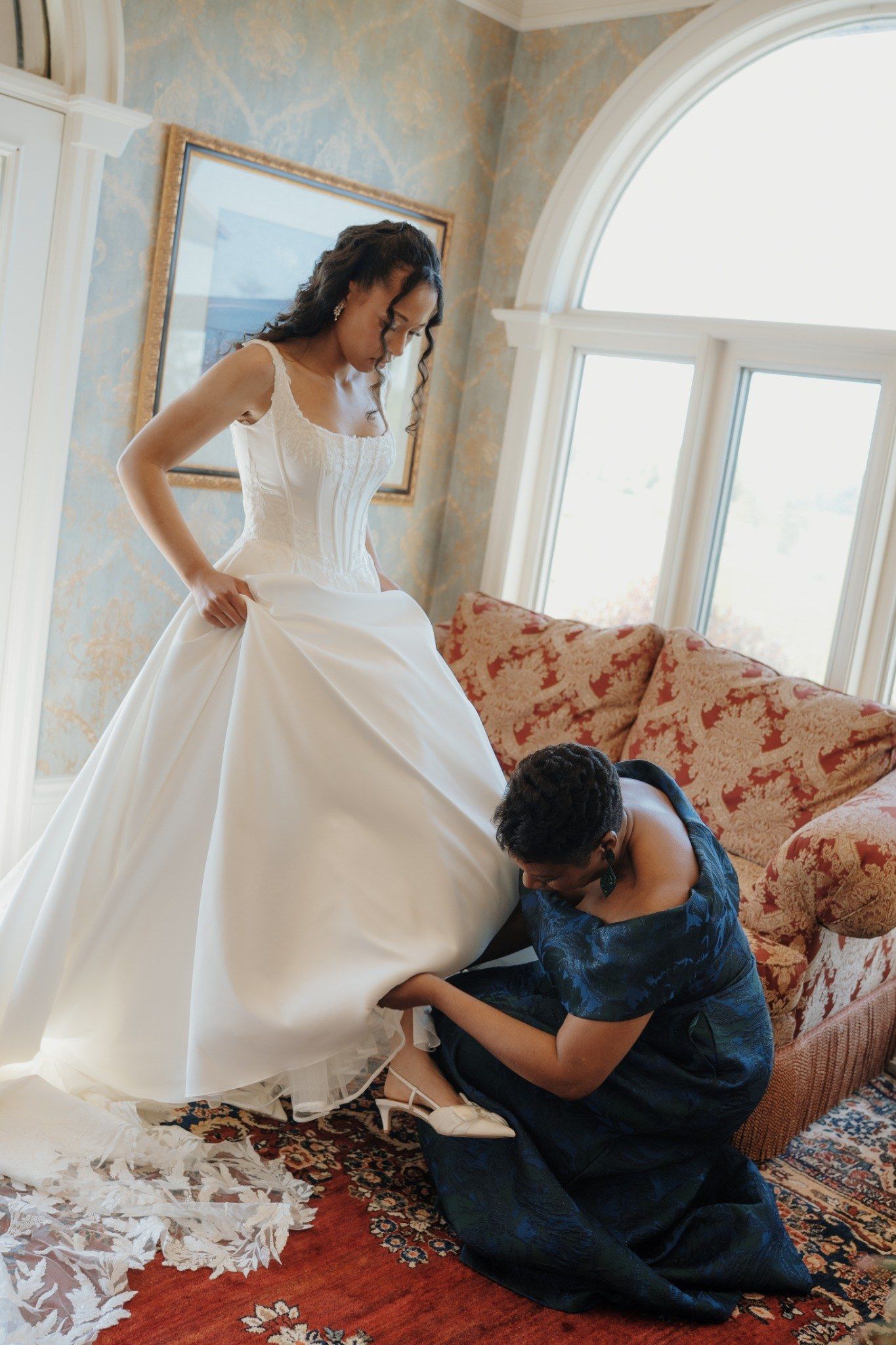 Bridesmaid in navy dress helps bride with shoes in elegant room with arched windows and floral wallpaper.