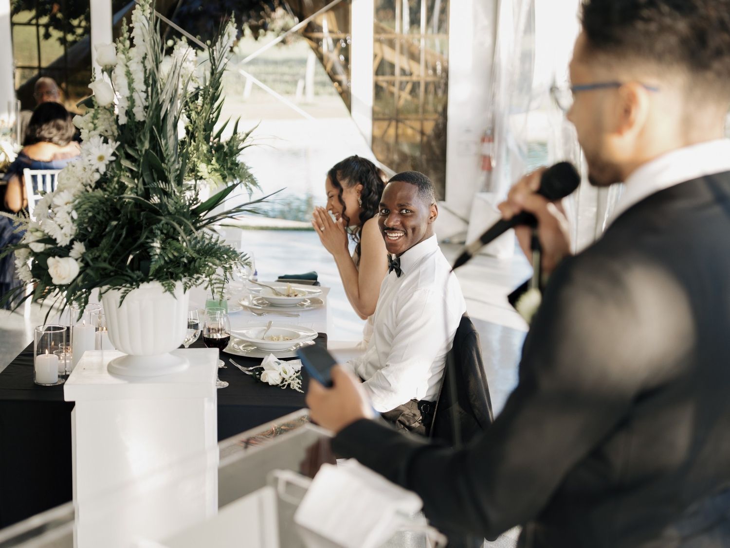 A joyful couple at their wedding reception laughs as a groomsman speaks into a microphone during a toast.