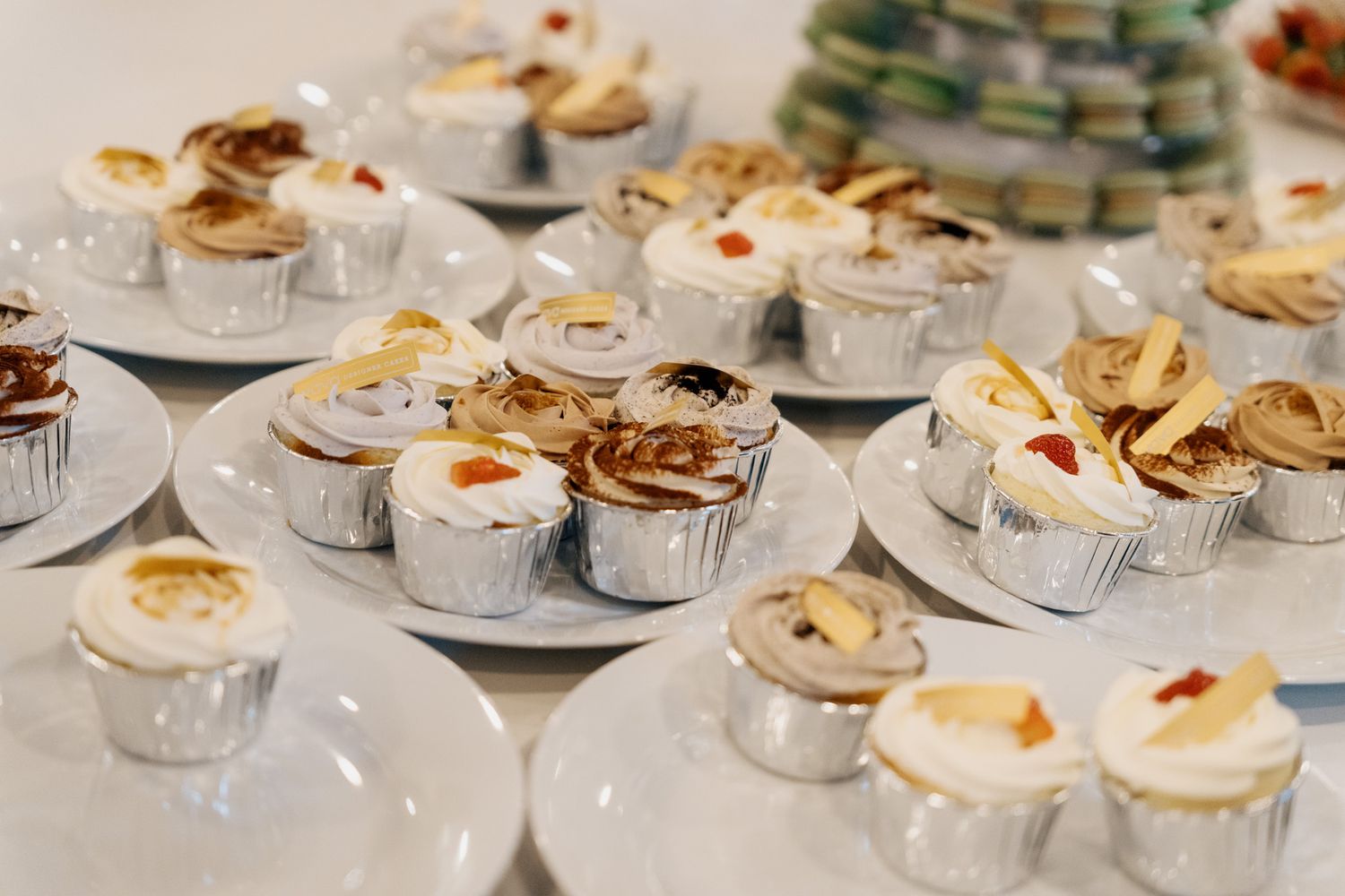 Assorted decorated cupcakes in silver foil liners on white plates at a Razi Estate forest wedding dessert table for candid reception photo