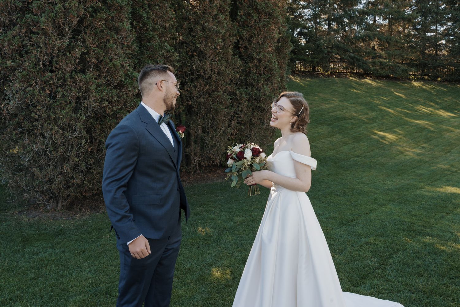 Bride in white off-shoulder gown holds bouquet while groom in navy suit smiles during outdoor first look.