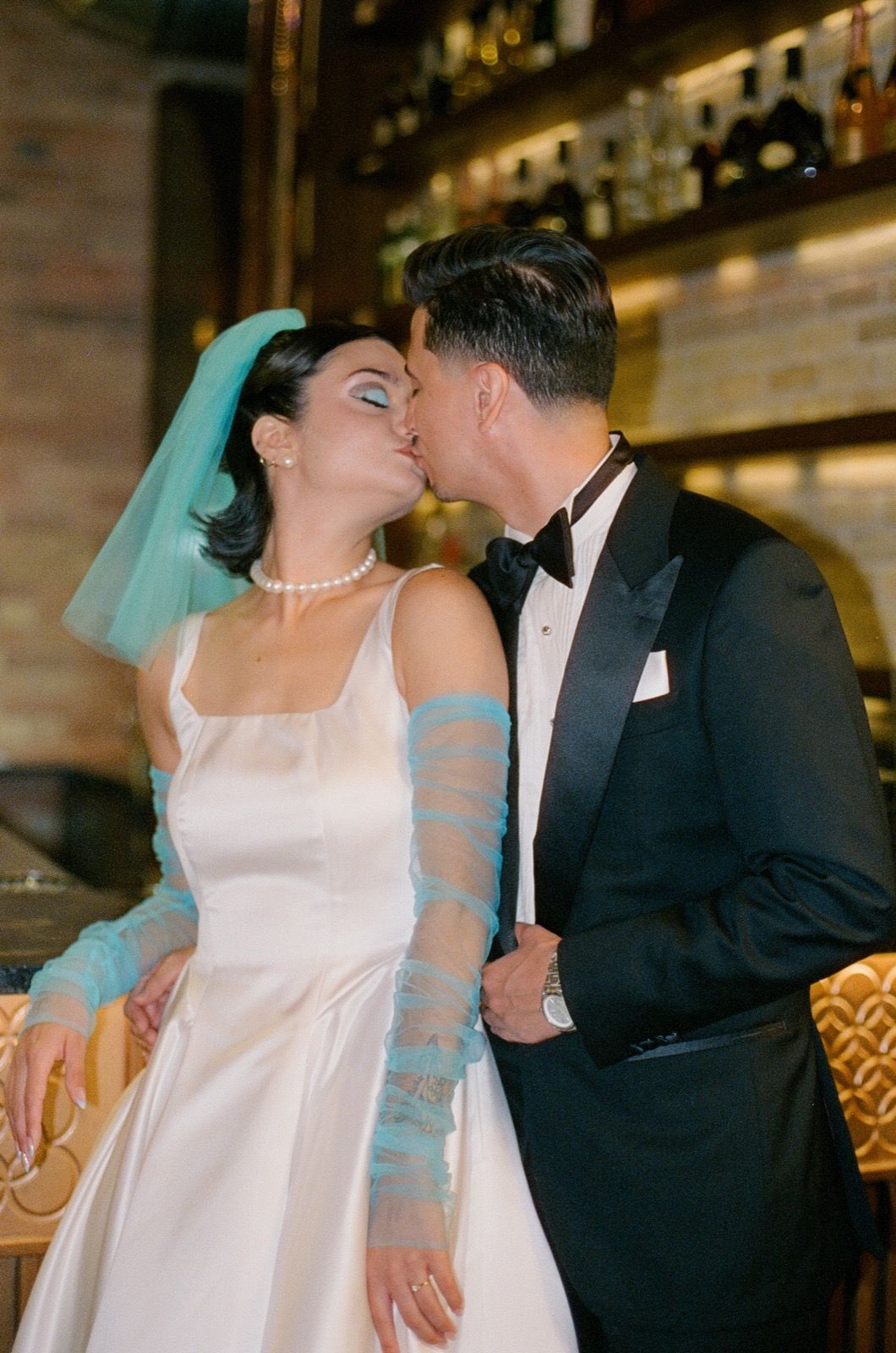 Bride and groom share a kiss, she wears a white gown with teal gloves and veil, he wears a classic black tuxedo.