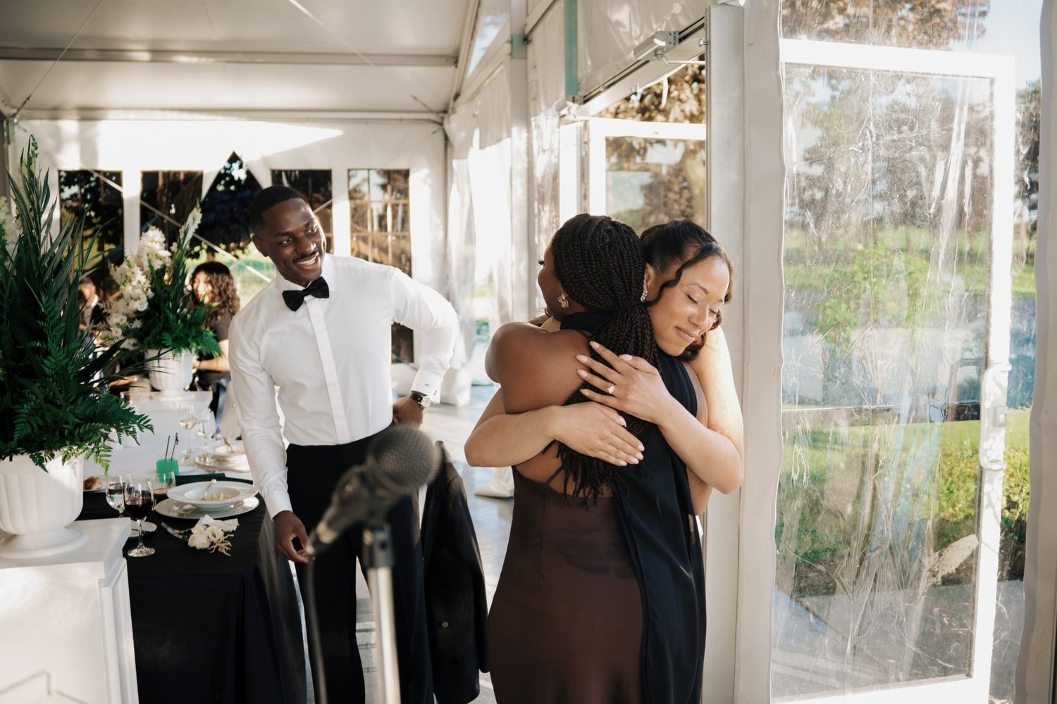 Two women embrace warmly at an elegant event while a smiling man in a tuxedo looks on inside a tent venue.