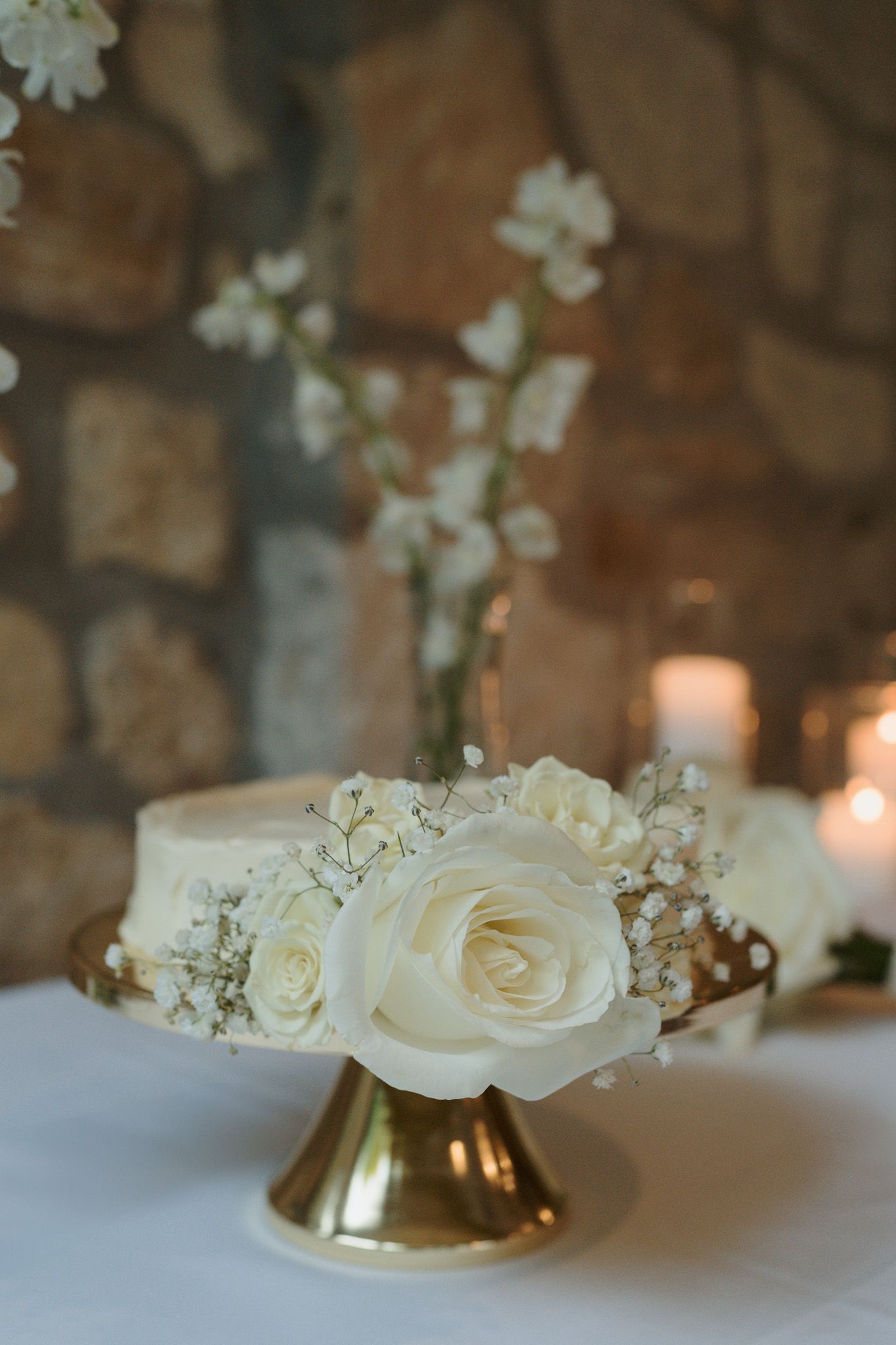 Elegant White rose wedding cake on a gold stand against a stone wall at Cambridge Mill, Ontario for editorial wedding photography
