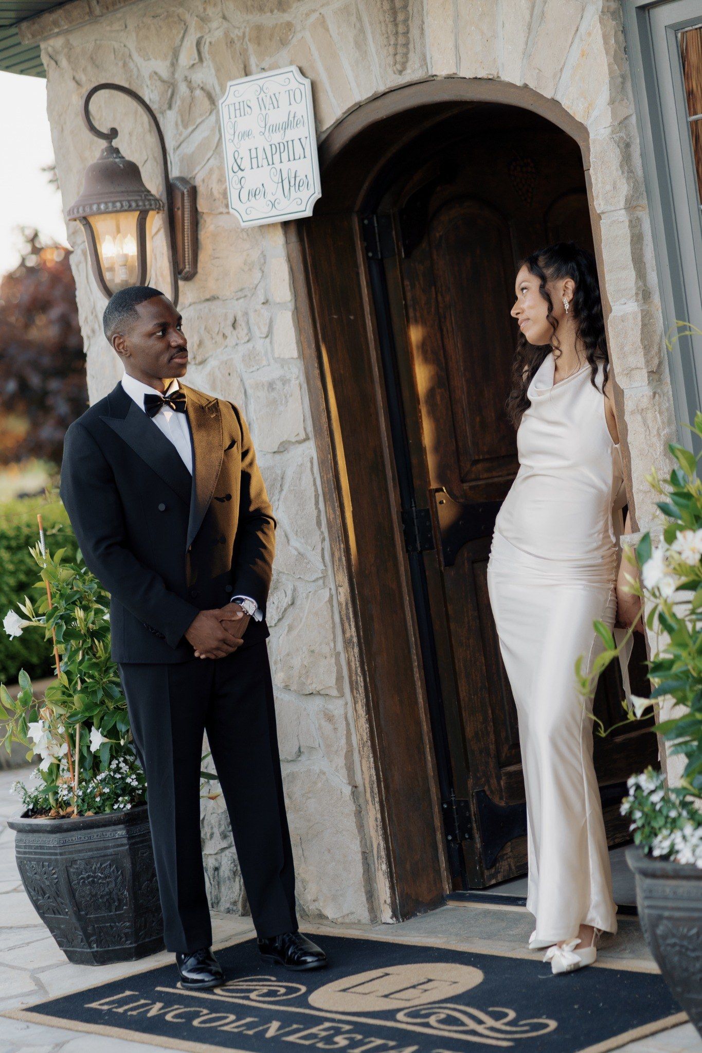 Elegant couple in wedding attire stand at stone venue entrance, groom in tuxedo and bride in white satin gown.