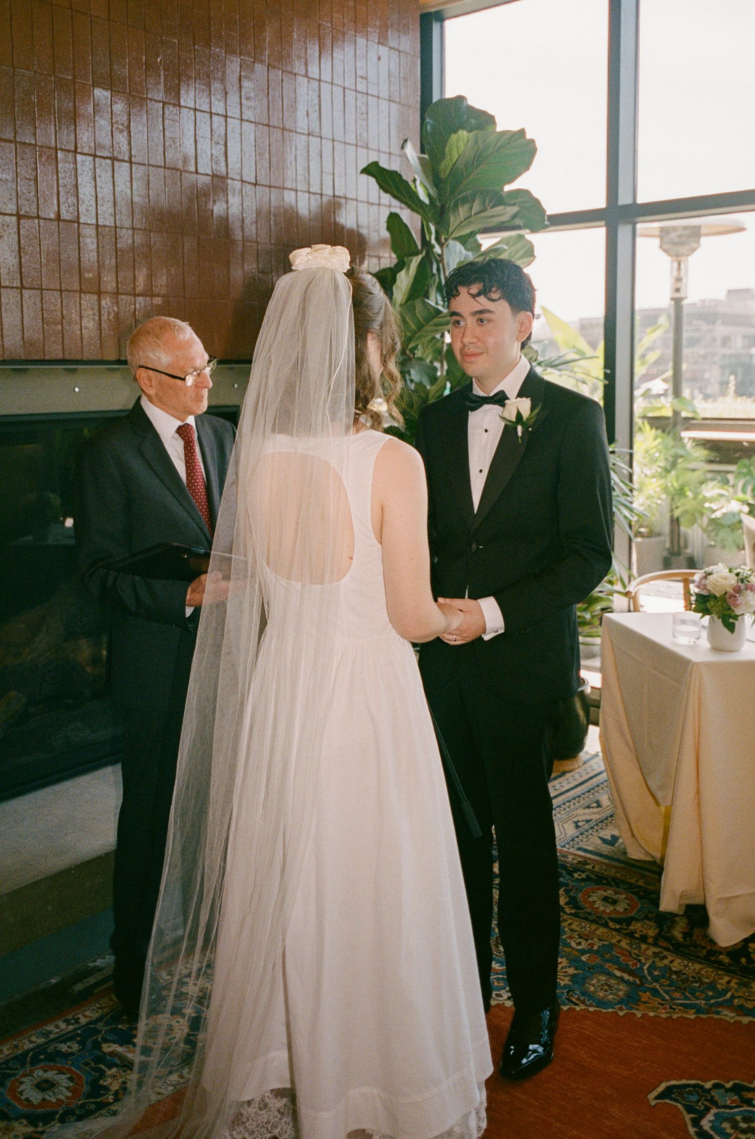 Bride in white gown and veil exchanges vows with groom in tuxedo during indoor wedding ceremony.