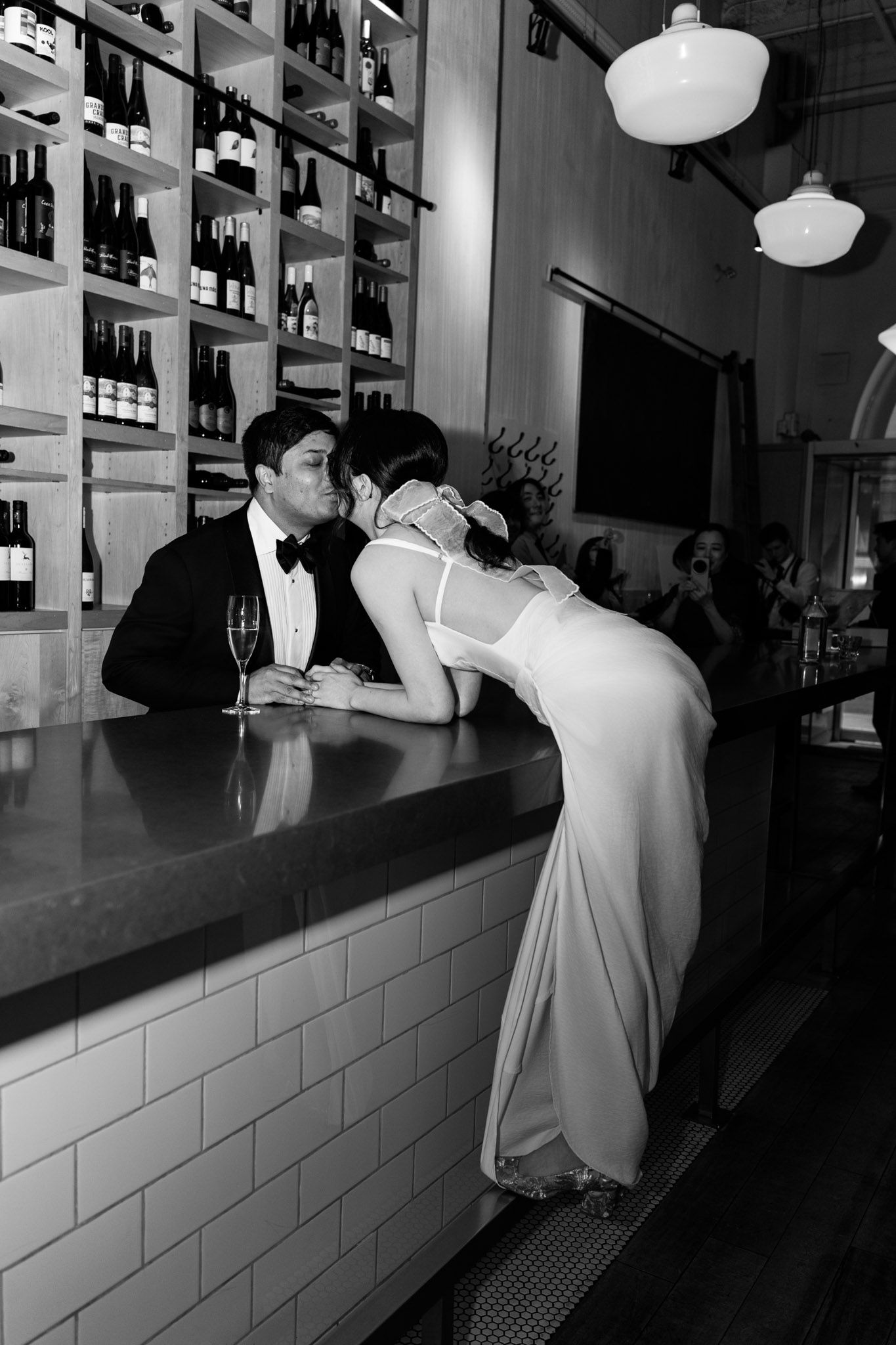 Black and white photo of a couple in wedding attire sharing a kiss at a wine bar counter with champagne.