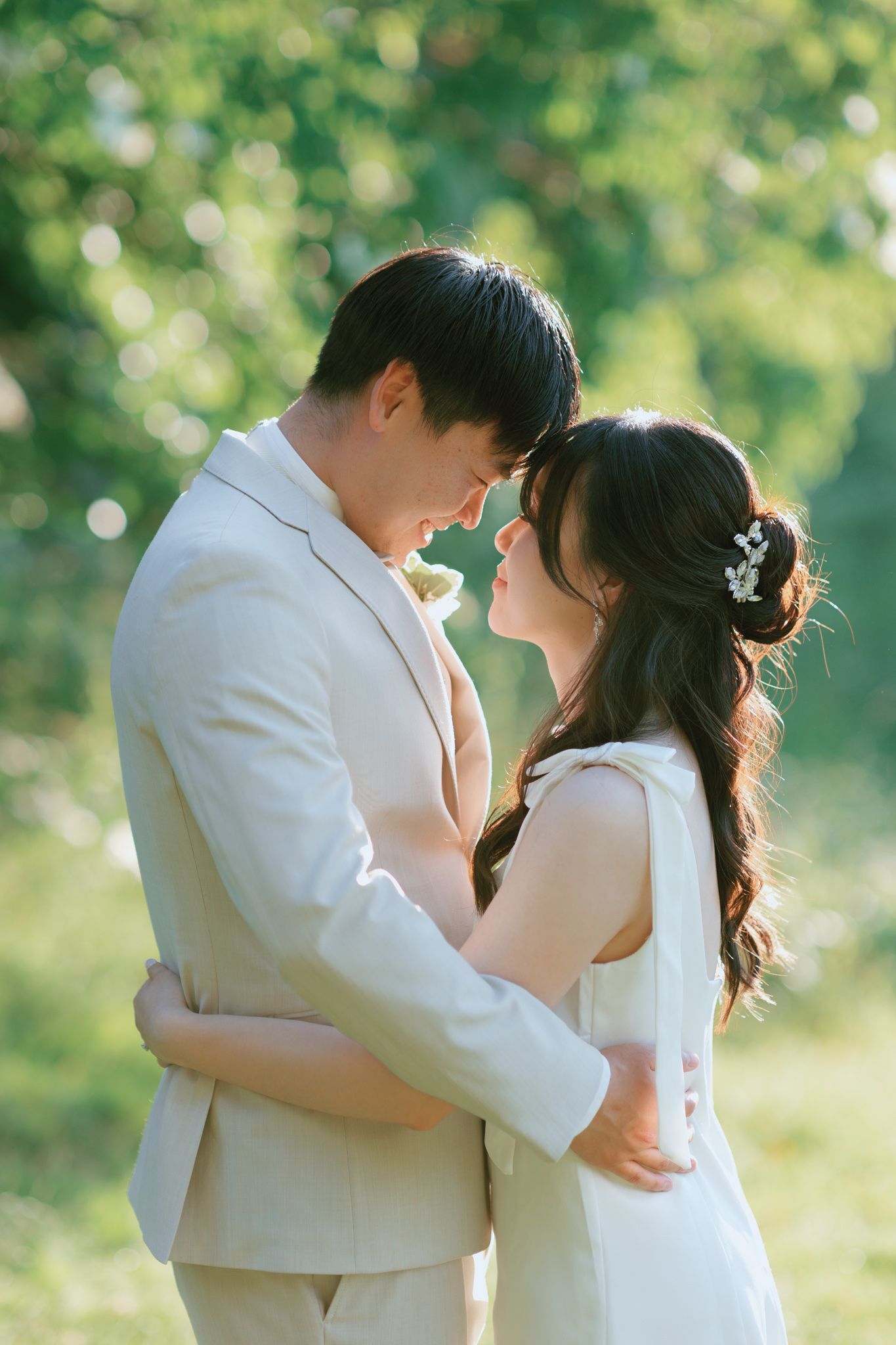 Couple in wedding attire embracing nose-to-nose in a sunlit green outdoor setting.