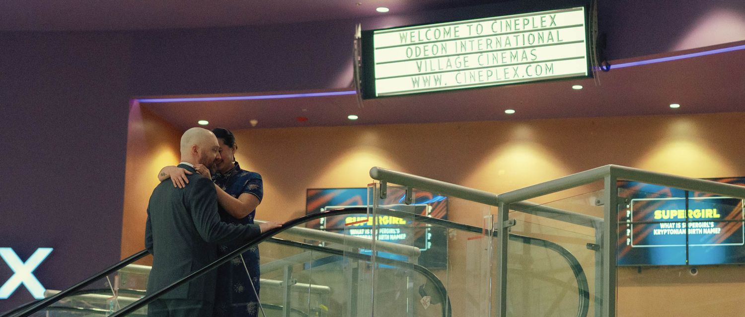 A couple embraces on an escalator inside a movie theater beneath a bright marquee sign.