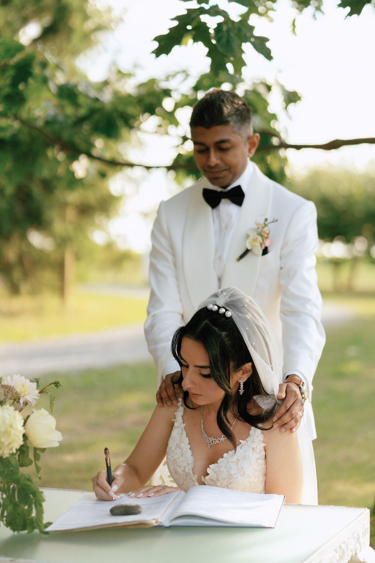 Bride signs wedding registry book outdoors as groom in white tuxedo with boutonniere stands behind her under green trees.