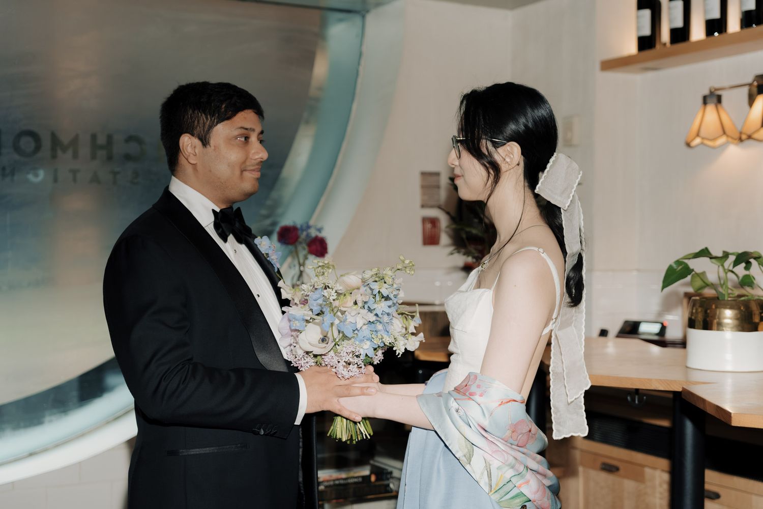Couple in wedding attire exchanging flowers indoors, groom in tuxedo and bride in white dress holding blue bouquet.