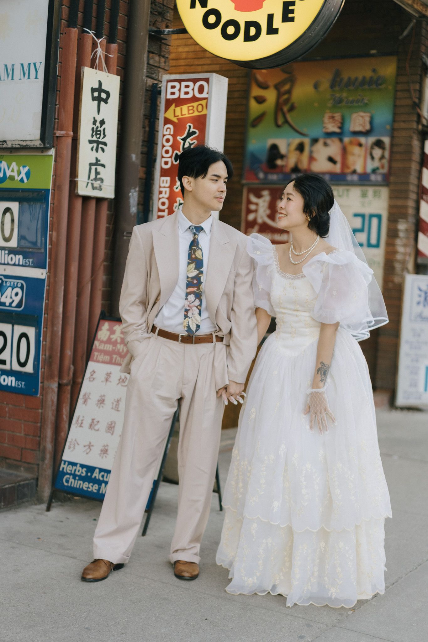 Couple in vintage wedding attire holding hands beneath neon Chinese storefronts during an 80s Hong Kong-inspired Toronto Chinatown shoot