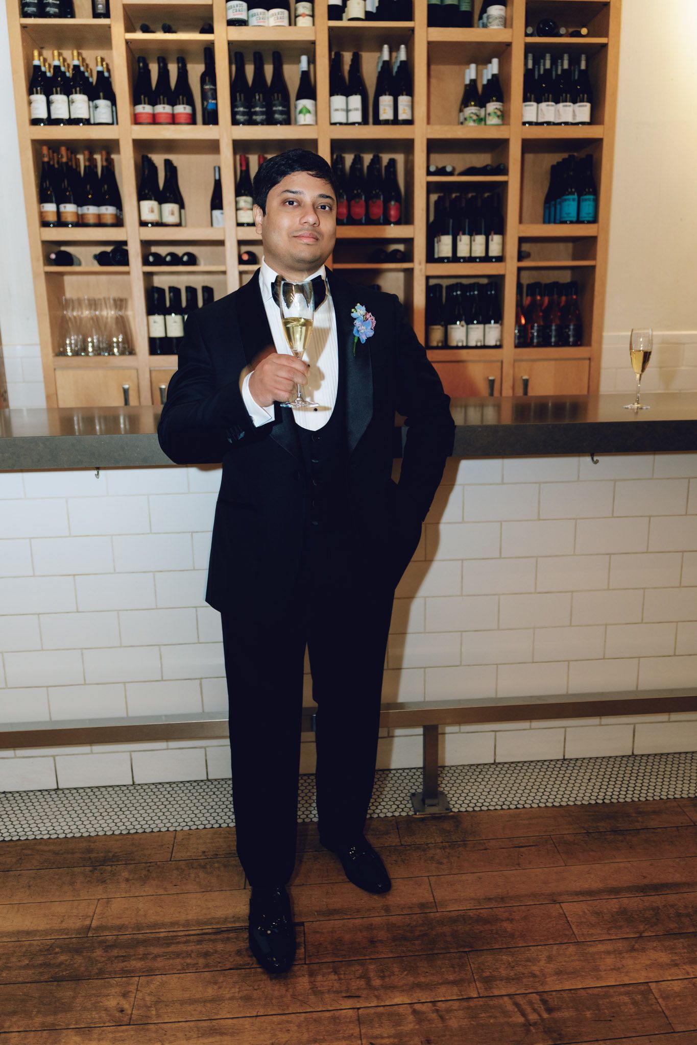 Man in black tuxedo holding champagne glass standing in front of wine bottle display shelves at bar.