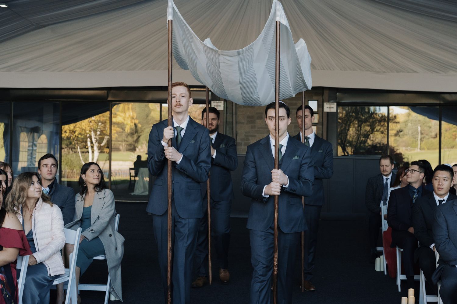 Groomsmen holding chuppah poles at The Manor in King City during an outdoor Jewish wedding ceremony with seated guests.