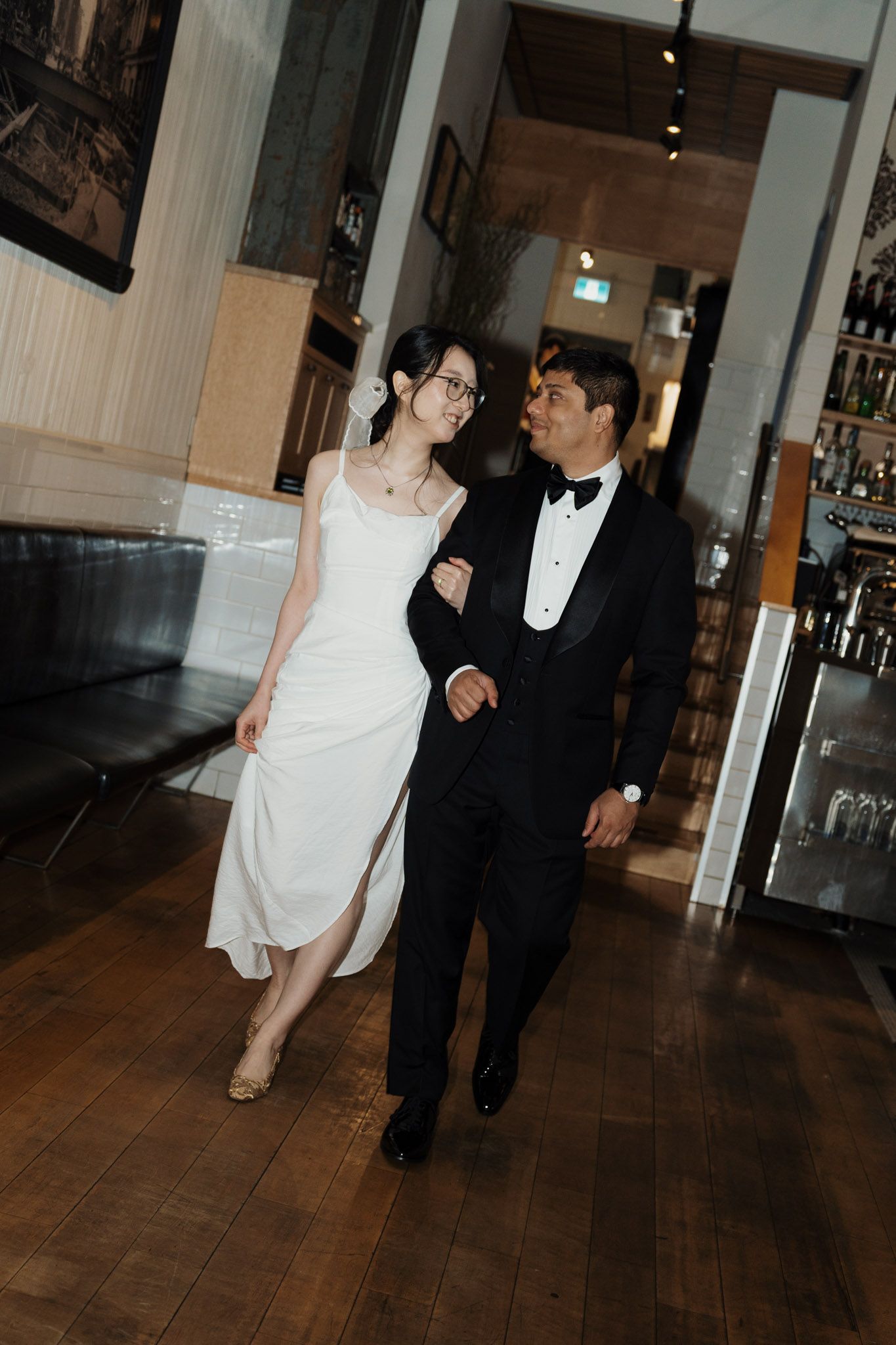 Bride in white gown and groom in black tuxedo walk arm in arm through an elegant restaurant venue.