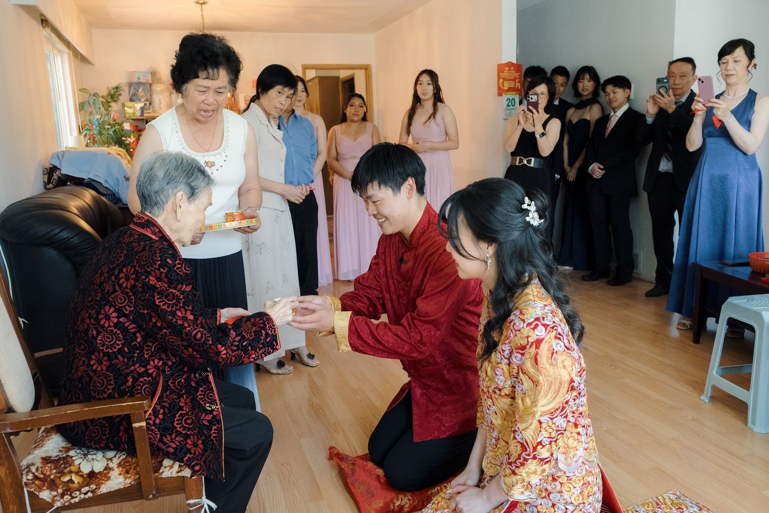 Couple kneels before elderly family member during traditional Chinese tea ceremony at home wedding.