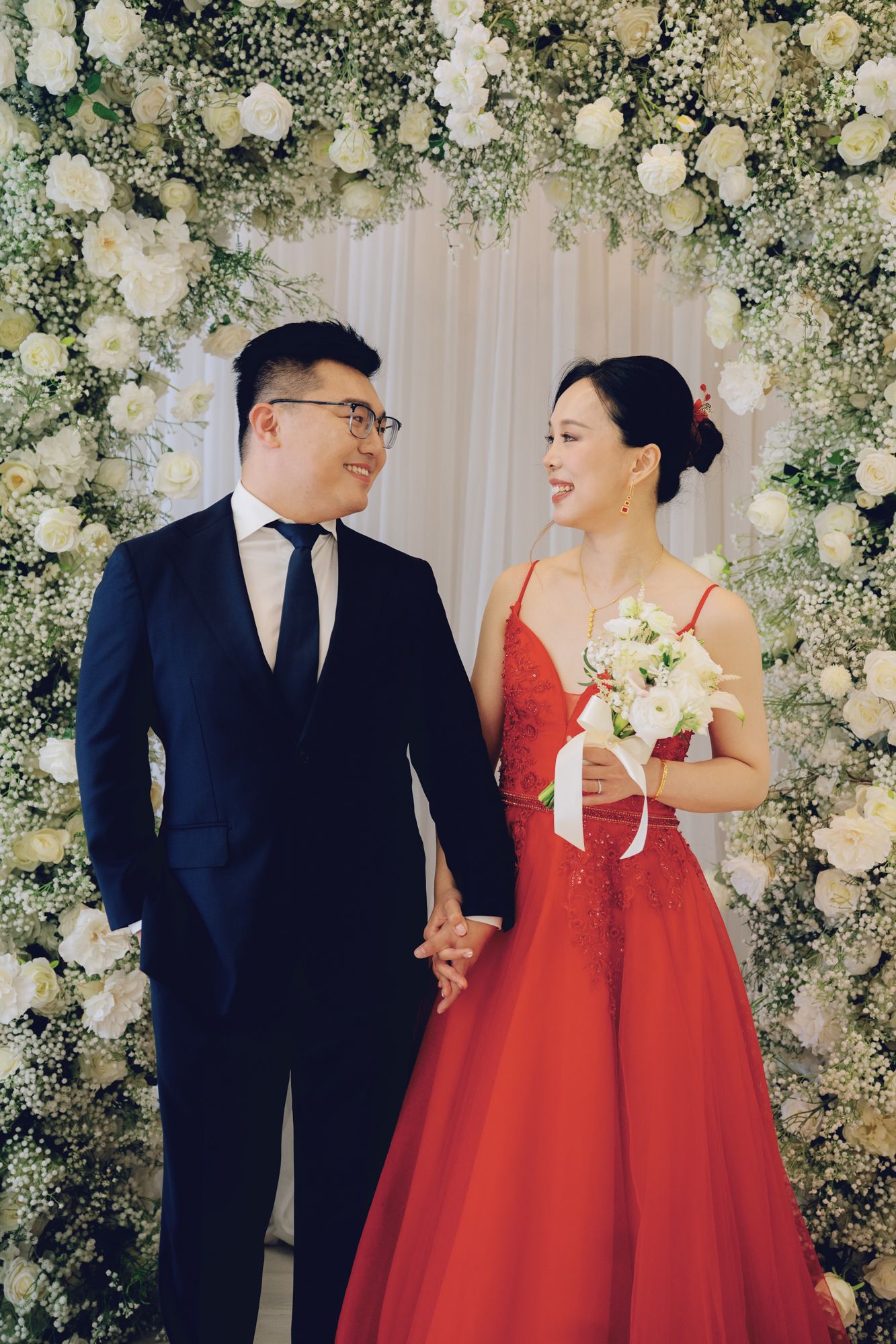 Bride in red gown and groom in navy suit holding hands beneath a floral arch at Bellamy Loft in North York, Toronto.