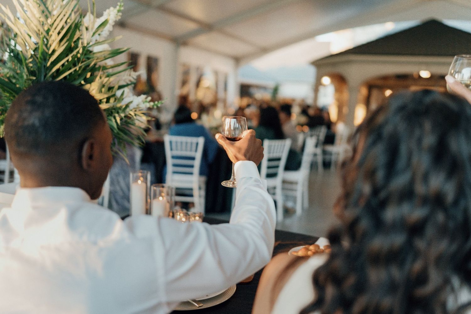 Couple raising wine glasses in a toast at an elegant outdoor wedding reception with white chairs and floral decor.