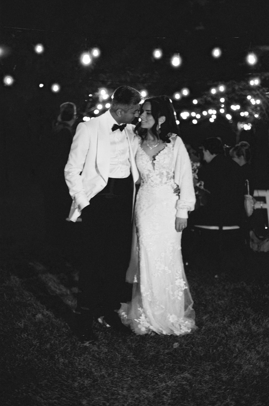 Black and white photo of a bride and groom sharing a romantic moment under string lights at an outdoor evening wedding reception.