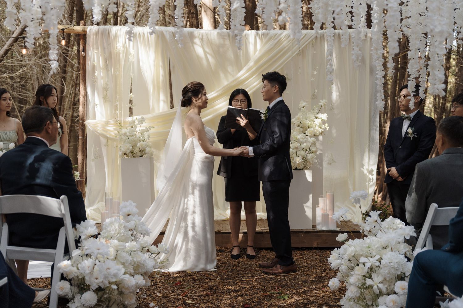 Couple exchanging vows beneath a white floral arch and draped fabric at Razi Estate in Schomberg for forest wedding photography.