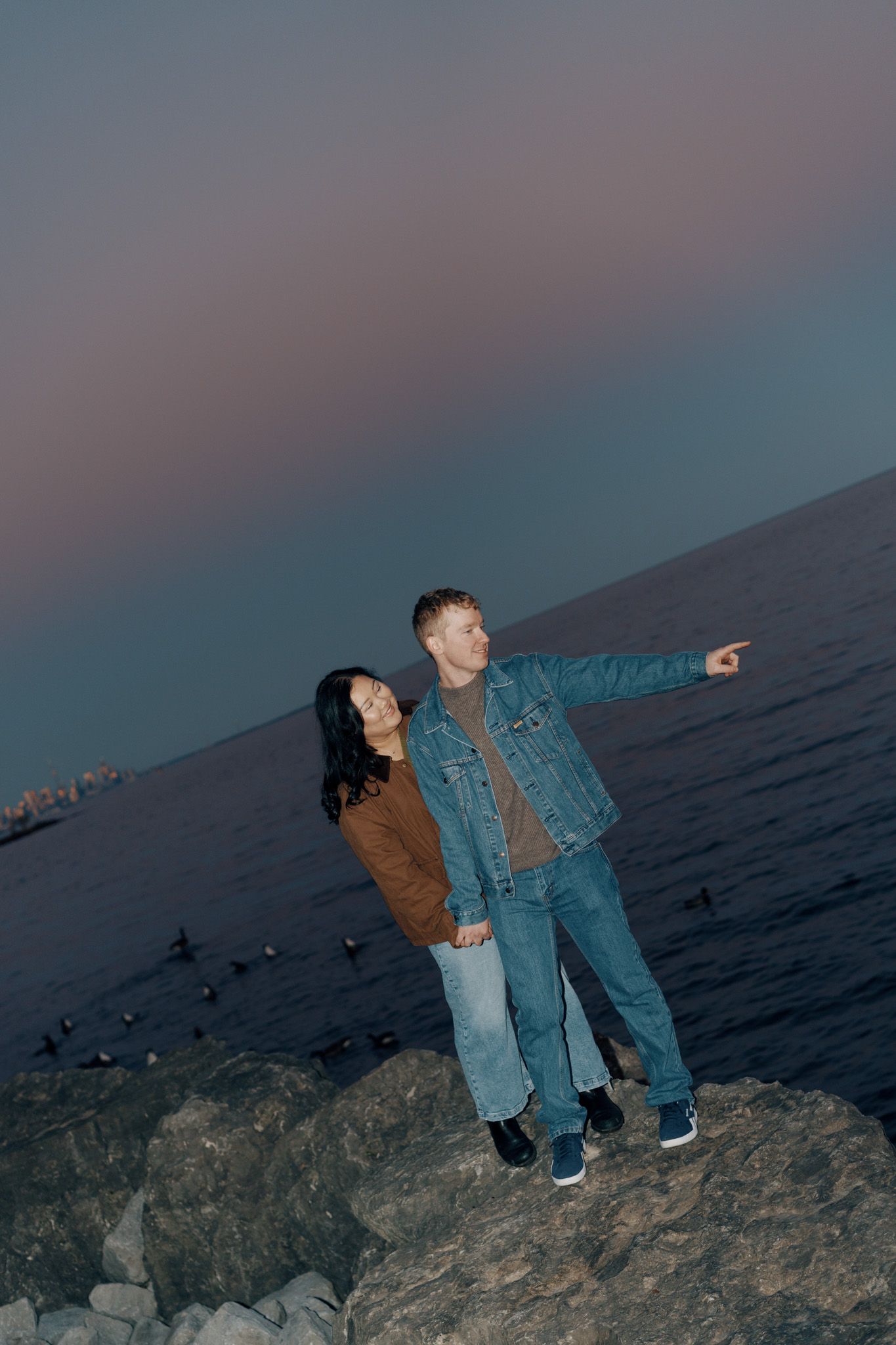Couple standing on lakeside rocks at dusk during a Mississauga beach sunset portrait session with cinematic engagement photos.