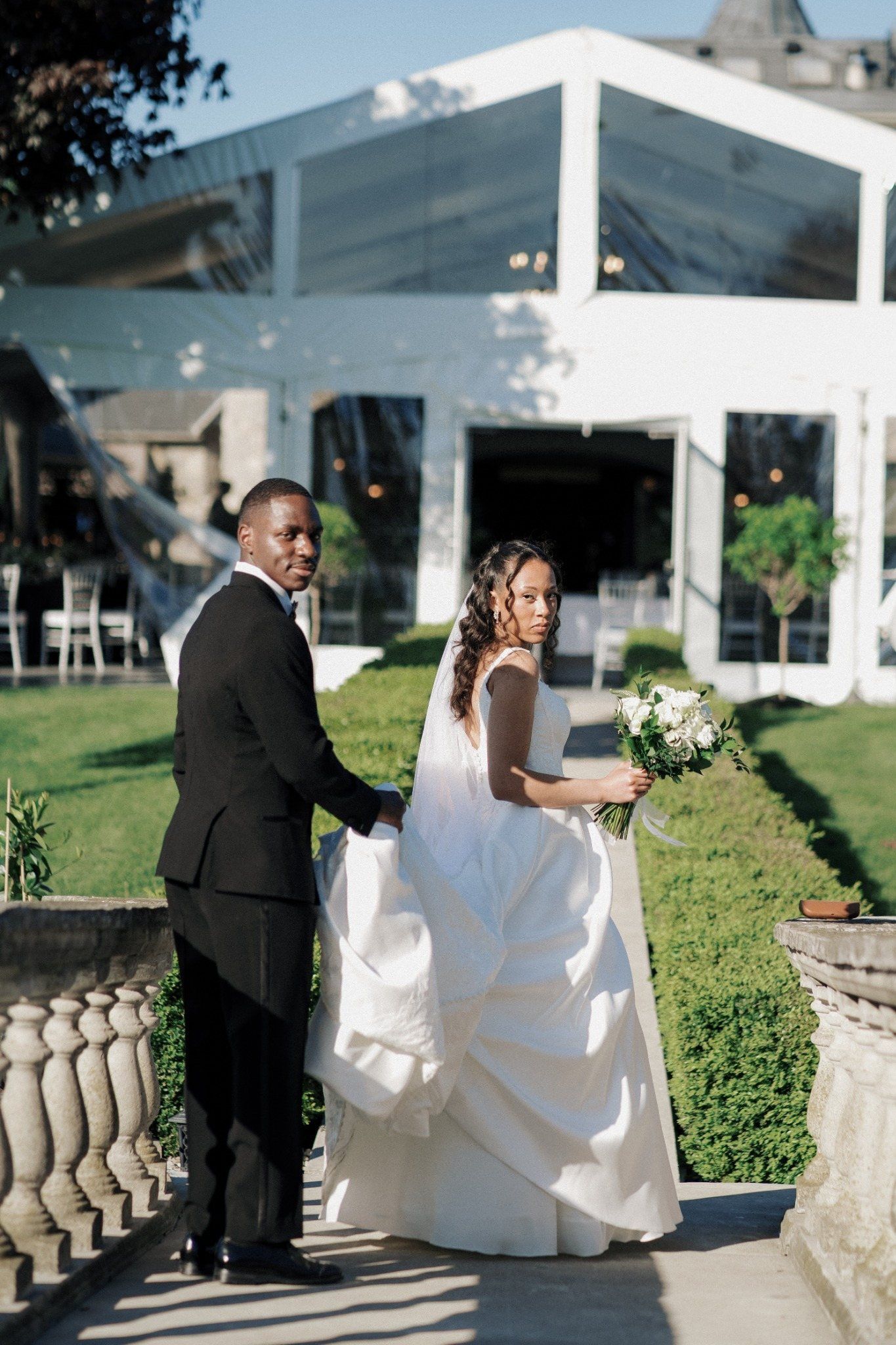 Elegant Black couple in wedding attire holding hands outside a white tent venue, bride holding white bouquet.